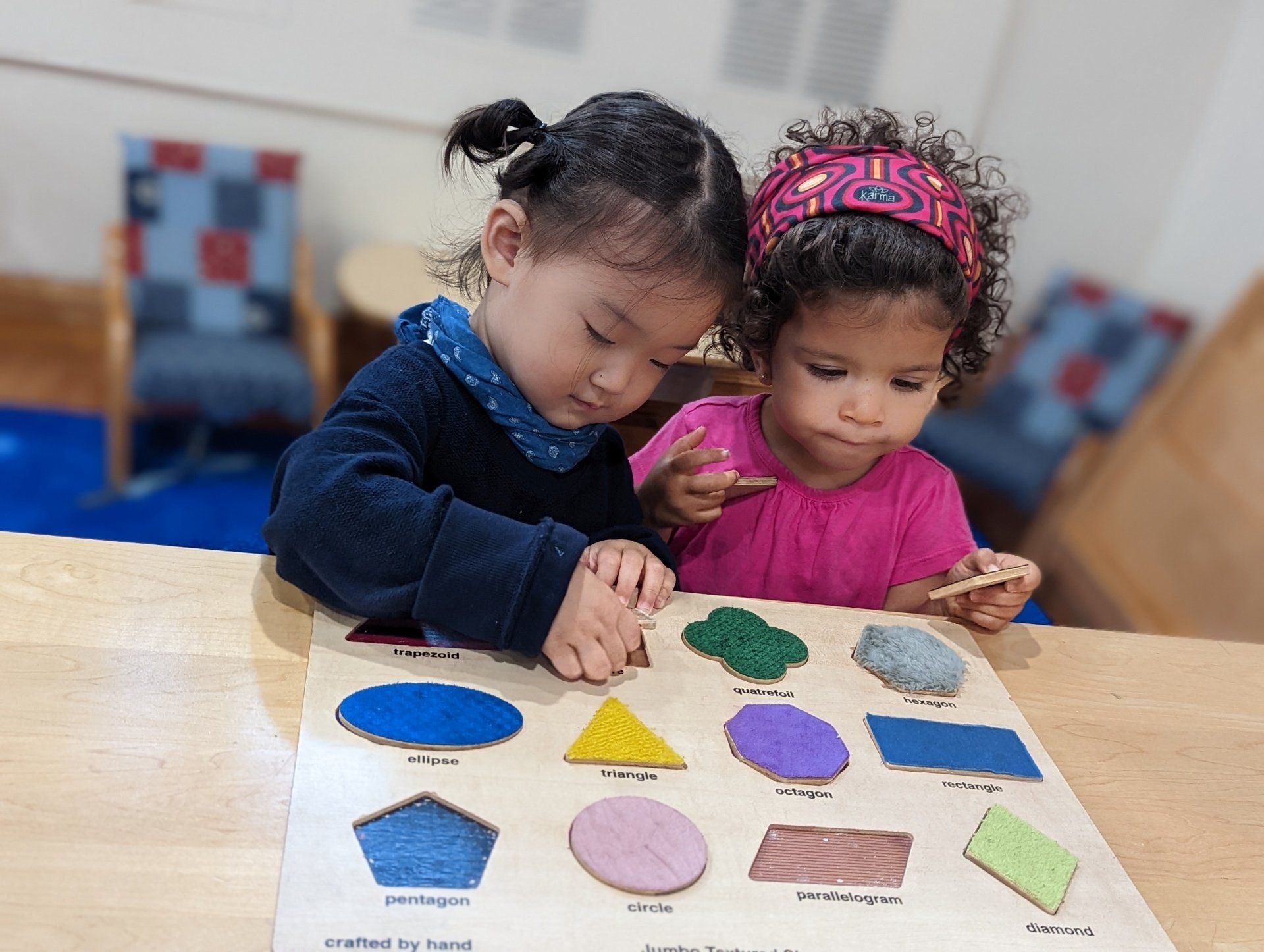 Two young girls with puzzle, matching shapes on a wooden table. One has pigtails, the other a headband.