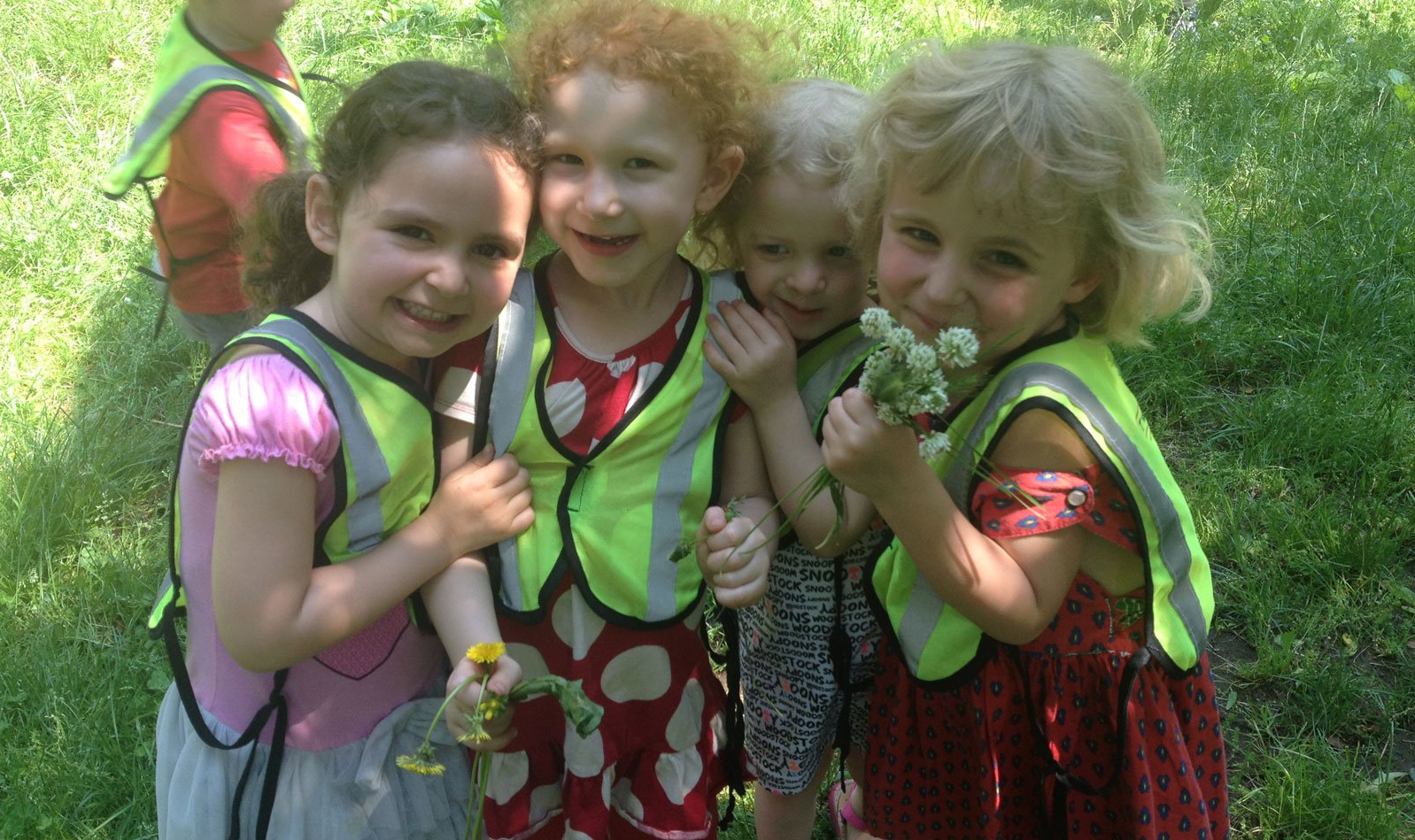 Montessori children in vests smile, gather in sunny green grass.
