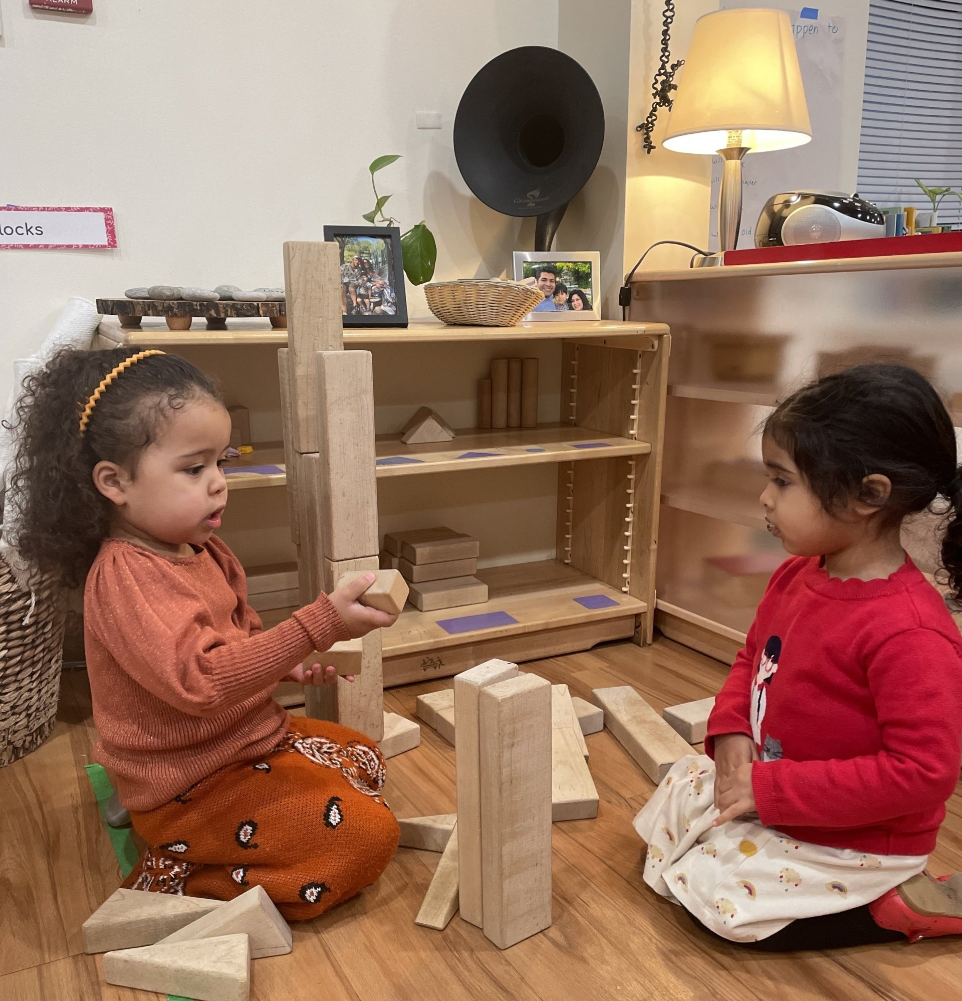 Two Montessori children are playing with wooden blocks indoors. One builds a tower, the other watches.