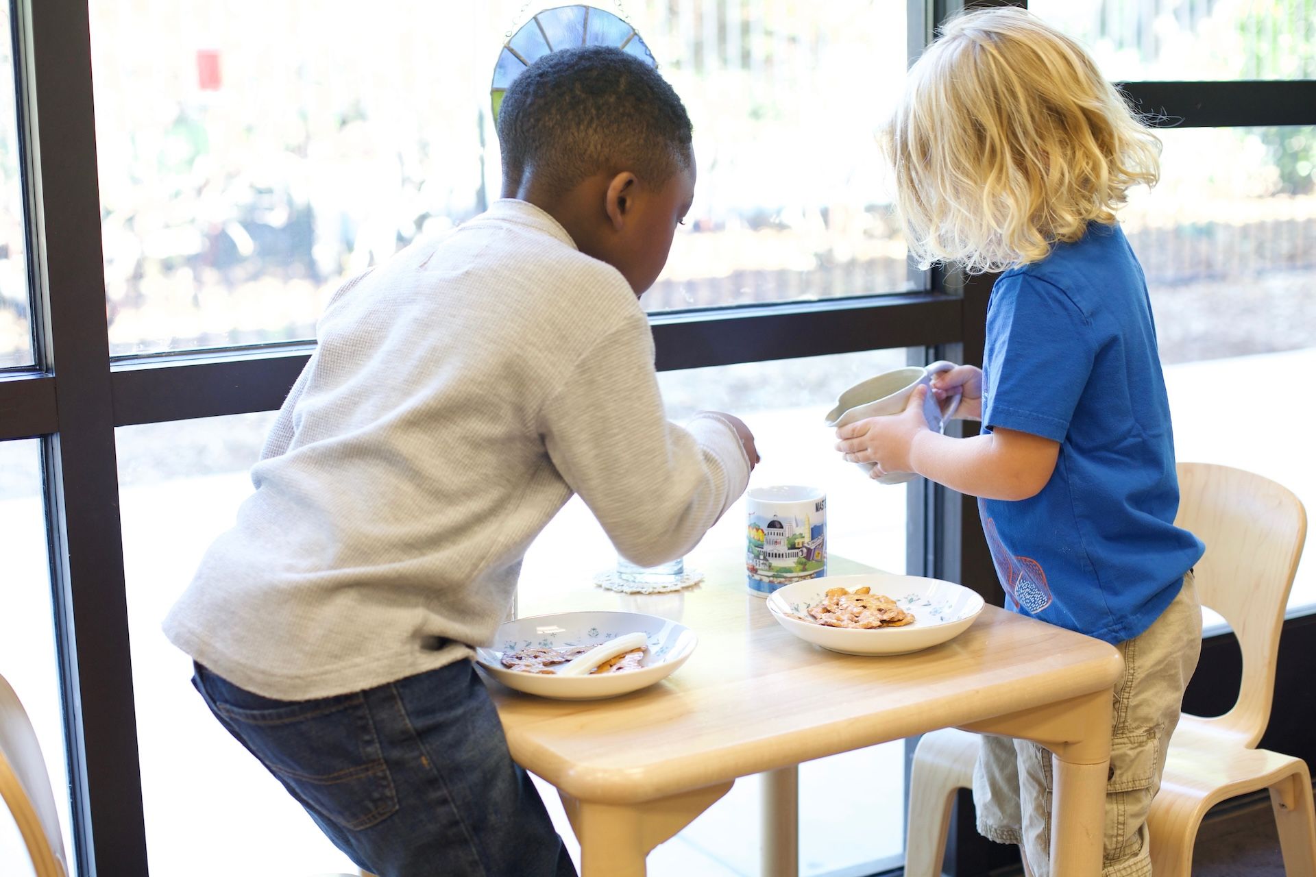 Two children at a table by a window, eating. One boy in gray, one girl in blue.