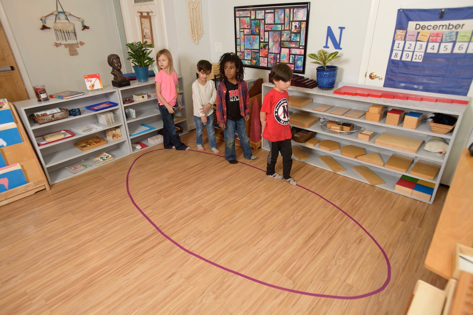 Children standing around a purple oval on a wooden floor in a classroom with shelves and learning materials.