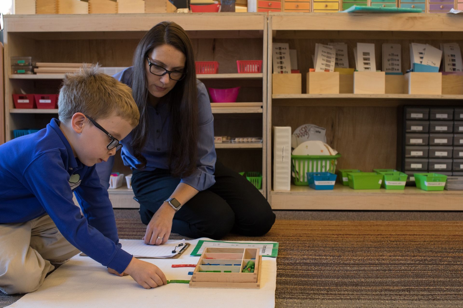A teacher helps a young boy with math on the floor of a classroom. Shelves in the background.