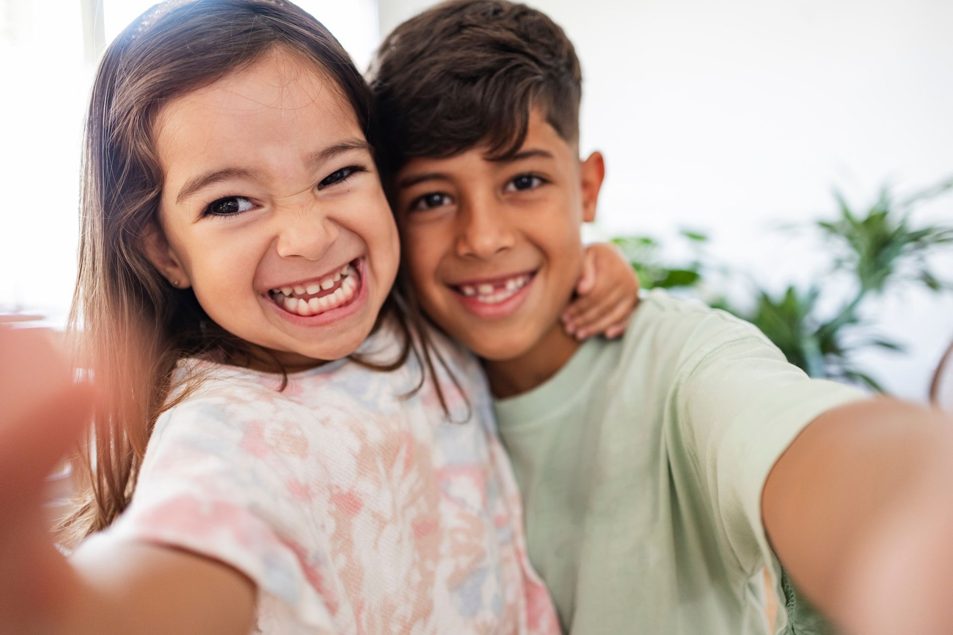 Two smiling children taking a selfie indoors. The girl has a big smile, and the boy has his arm around her.