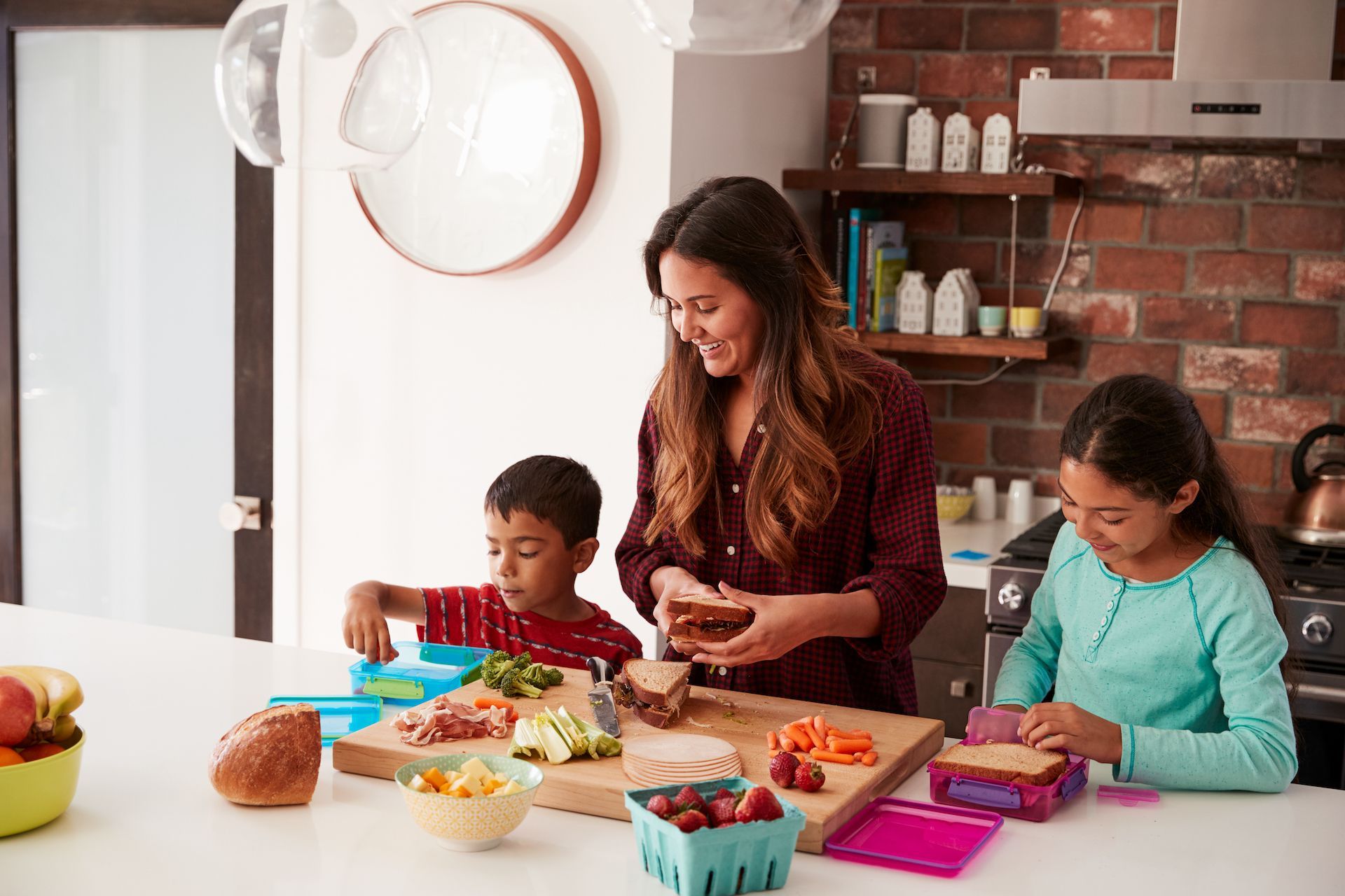 Woman and two children making lunches in a kitchen. They smile while preparing food on a white countertop.