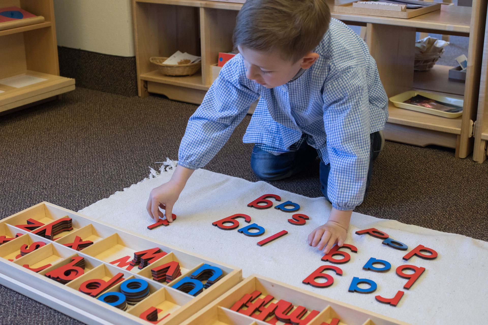 Young boy working with alphabet letters in a Montessori classroom; red and blue letters, wooden trays.