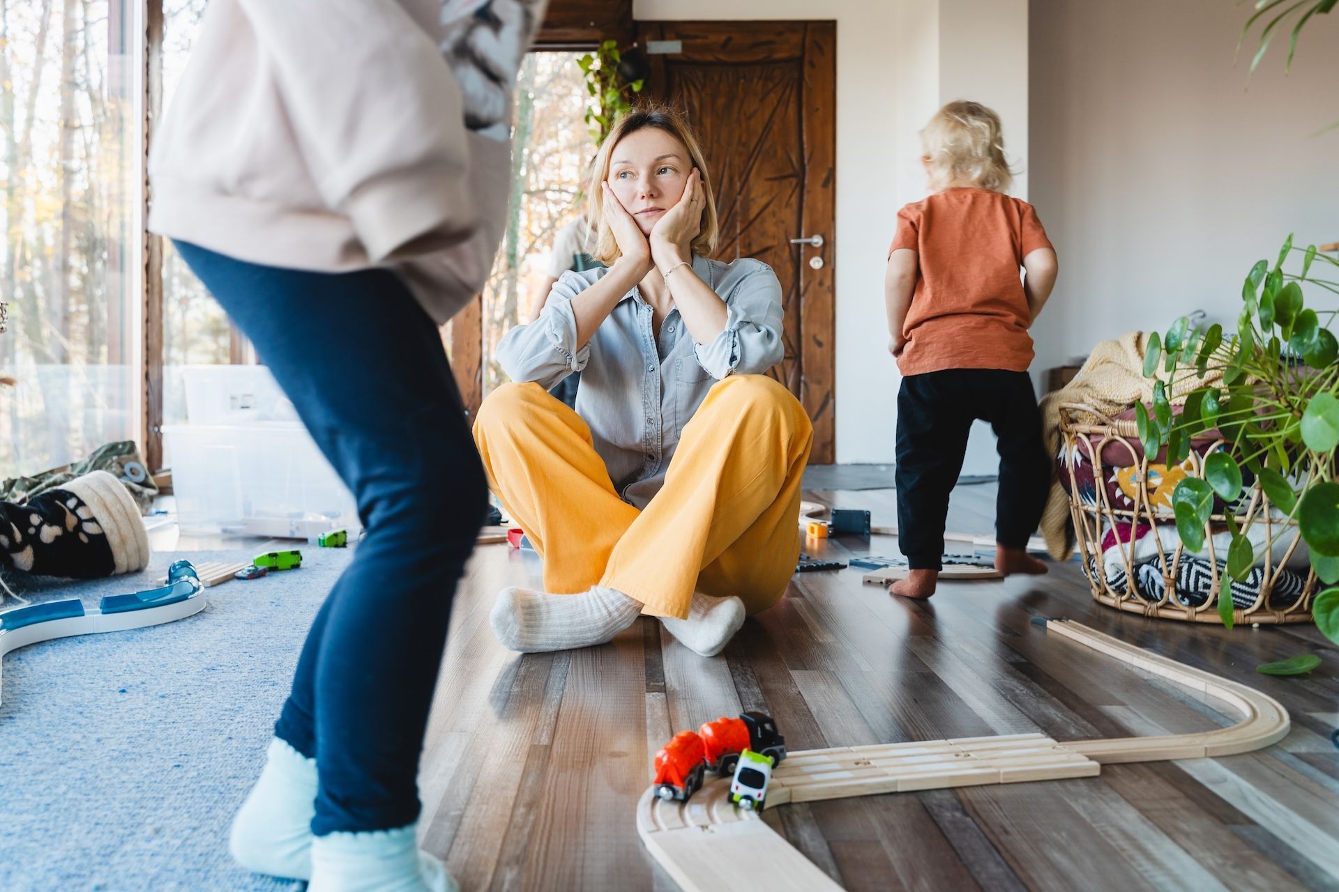 Woman with children sits on floor, looking stressed. Scattered toys, wooden train track.