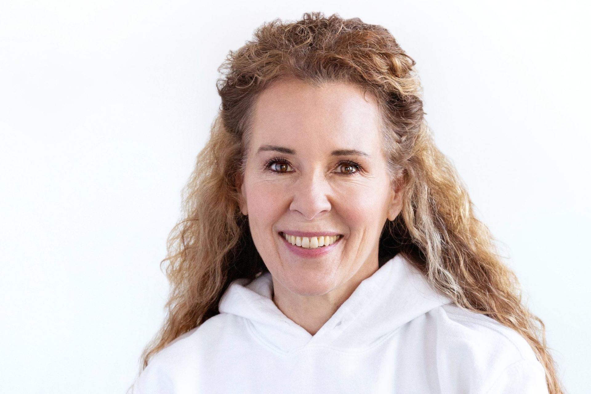 Woman with curly blonde hair smiles, wearing a white hoodie, against a white background.