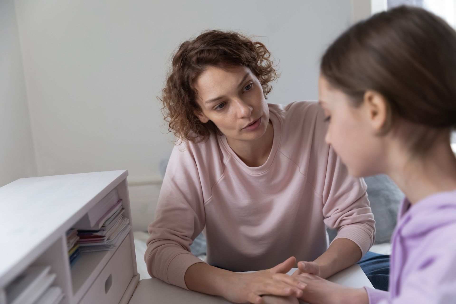 Woman comforts a girl, holding her hand in a room with a white desk and neutral walls.