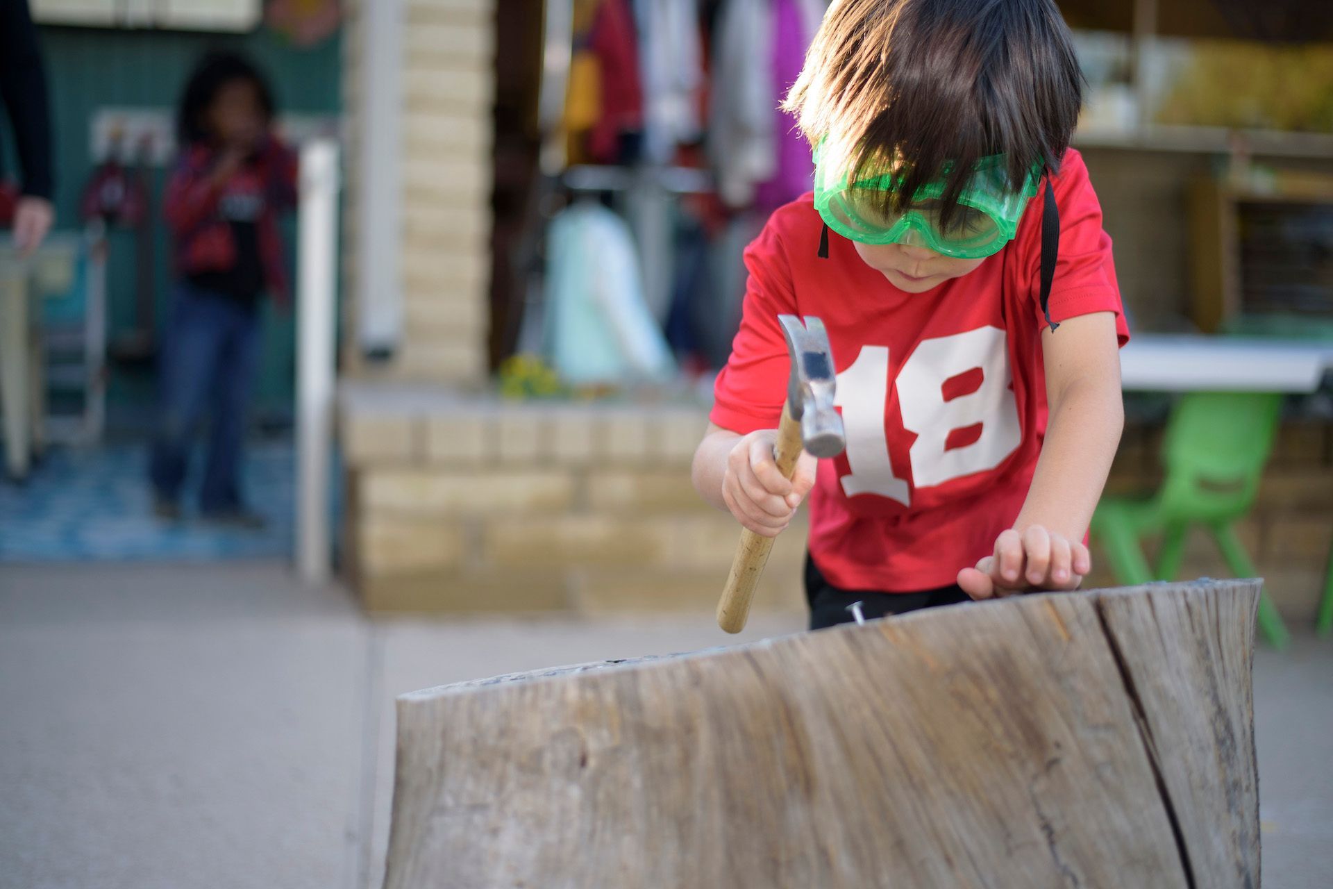 A young child in a red shirt and green headband hammers a piece of wood outdoors.