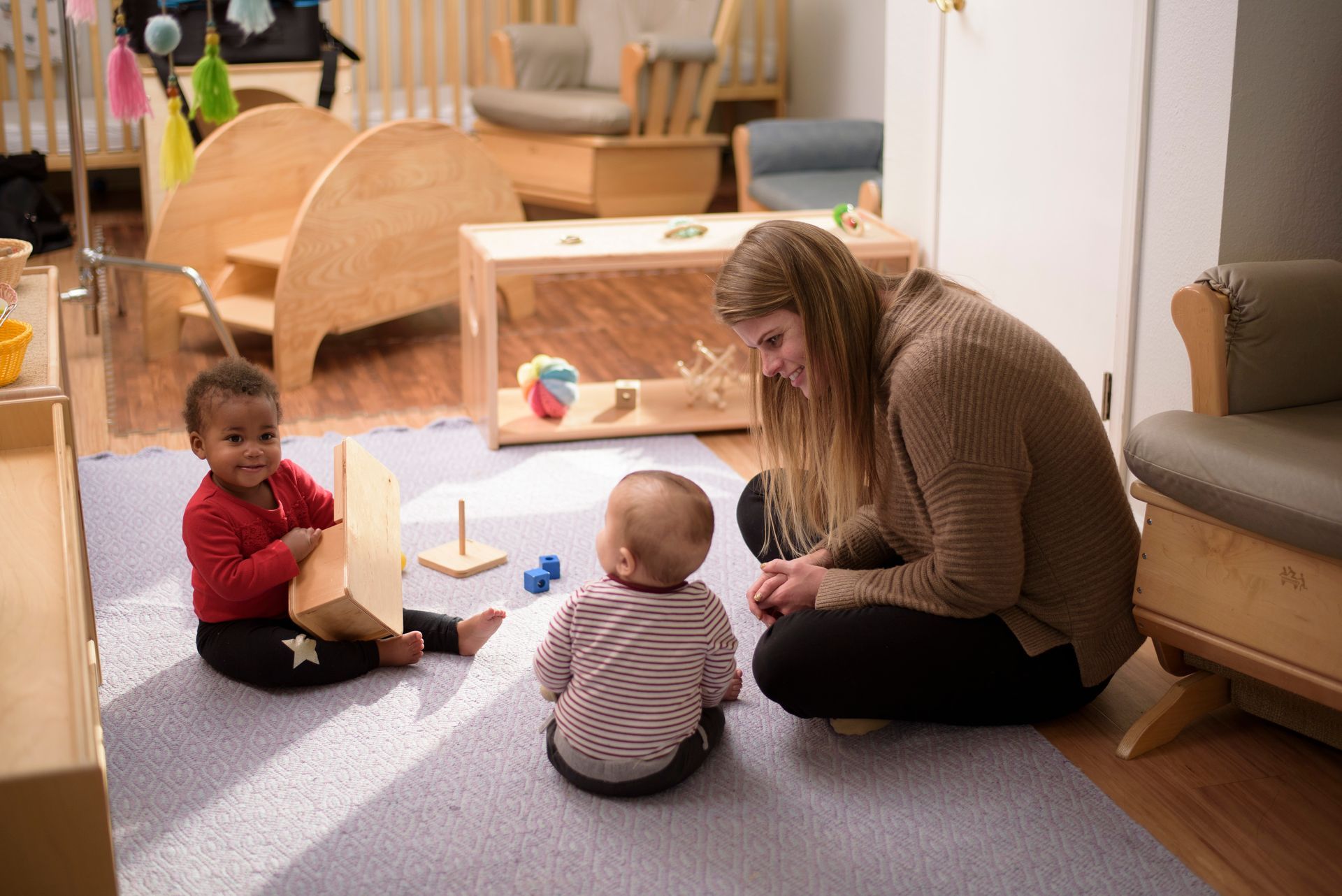 A Montessori guide is playing with two babies in a warmly lit playroom; babies are smiling, one holds a block.