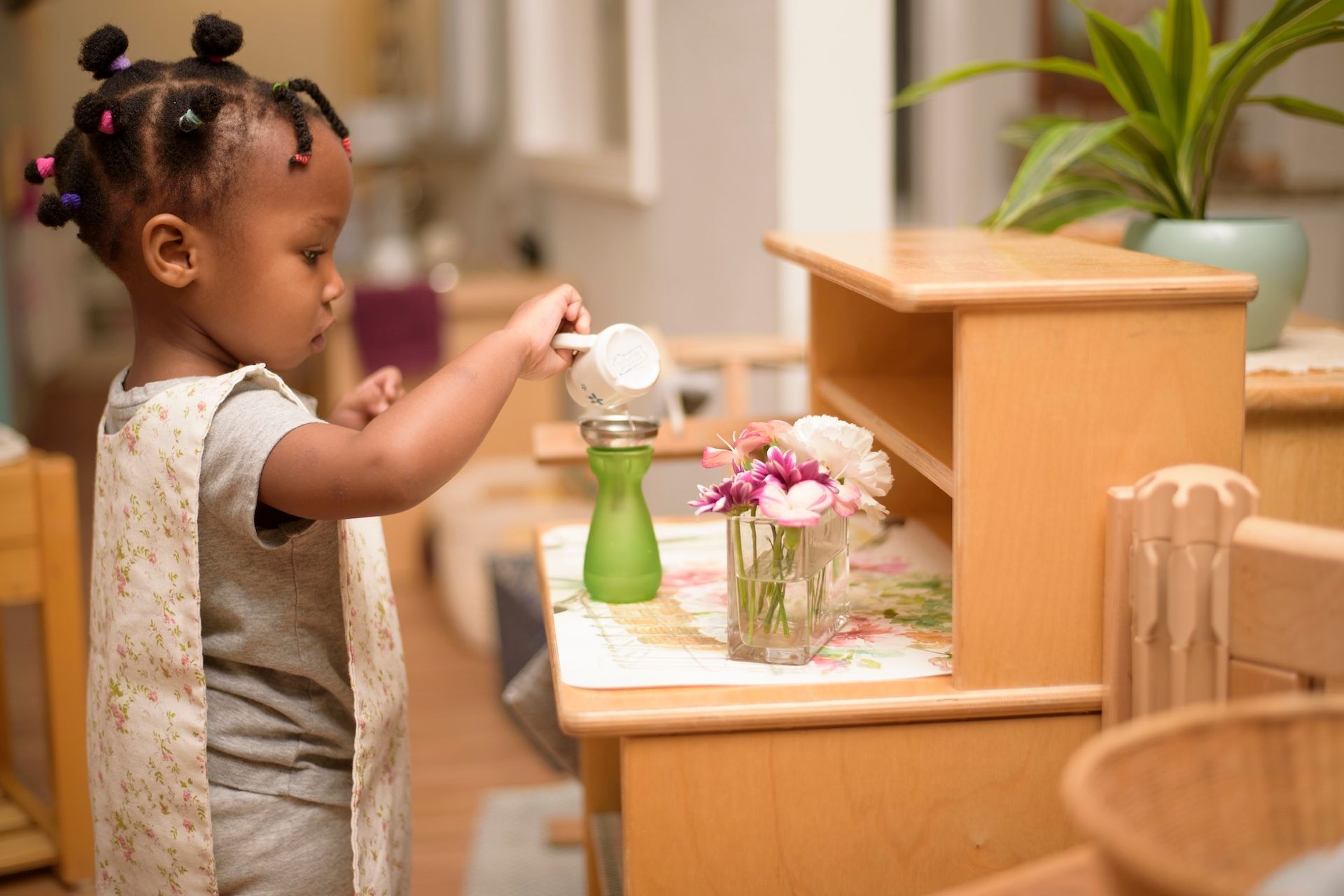 Young child pouring water from a pitcher onto a plant; indoors, wooden furniture.