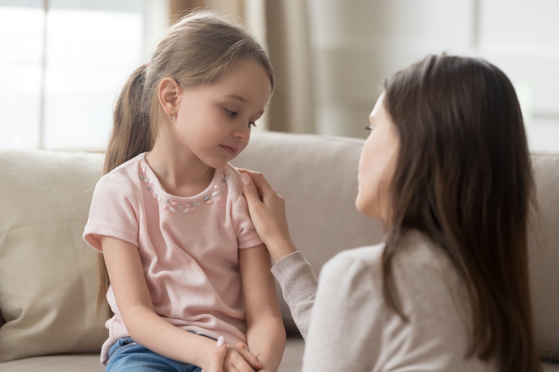Woman comforts a young girl with a hand on her shoulder, both looking down, indoors.