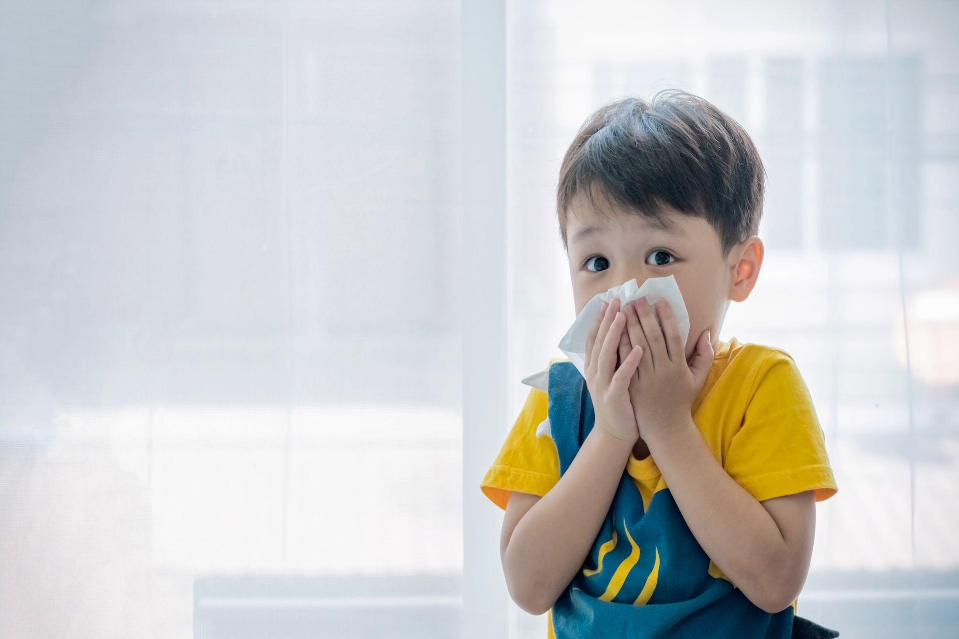 Boy blowing nose with tissue, looking surprised. Wearing yellow shirt, blue overall, indoors.