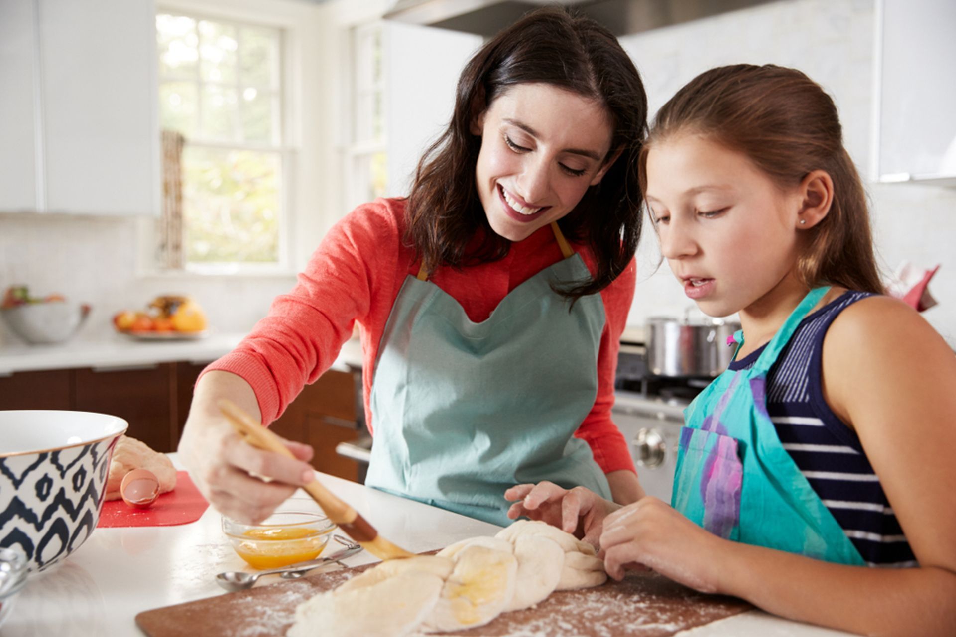 Woman and girl baking in a kitchen. Woman brushes dough with glaze as girl watches. Bright, smiling expressions.