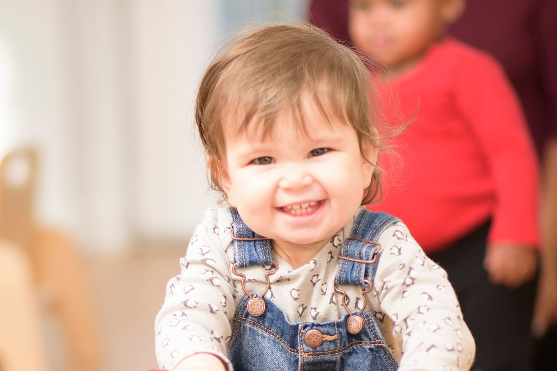 Smiling baby in denim overalls, indoors, another child in red shirt blurred in the background.