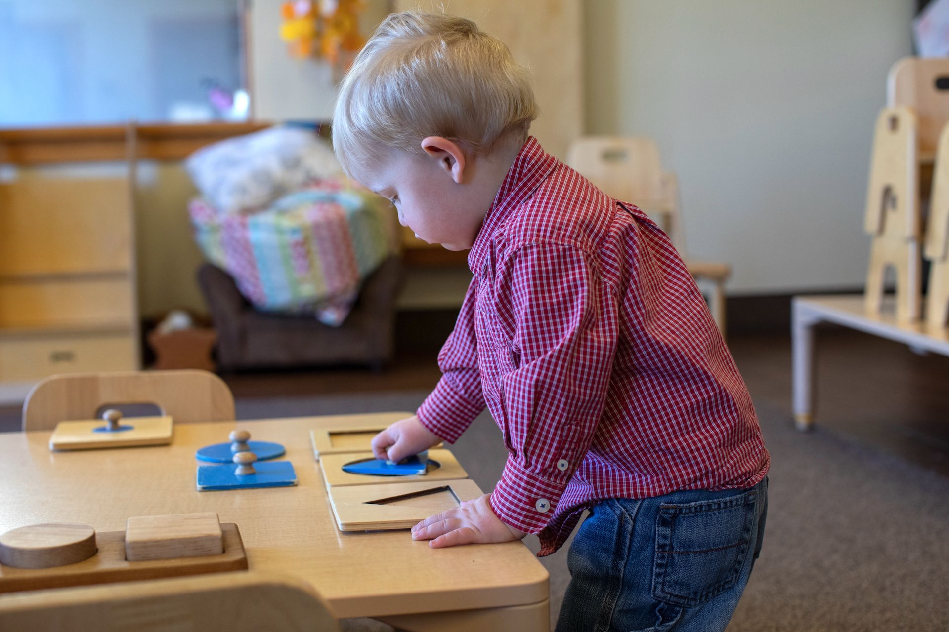 A young child, wearing a red shirt and jeans, works with a wooden puzzle at a table in a classroom.