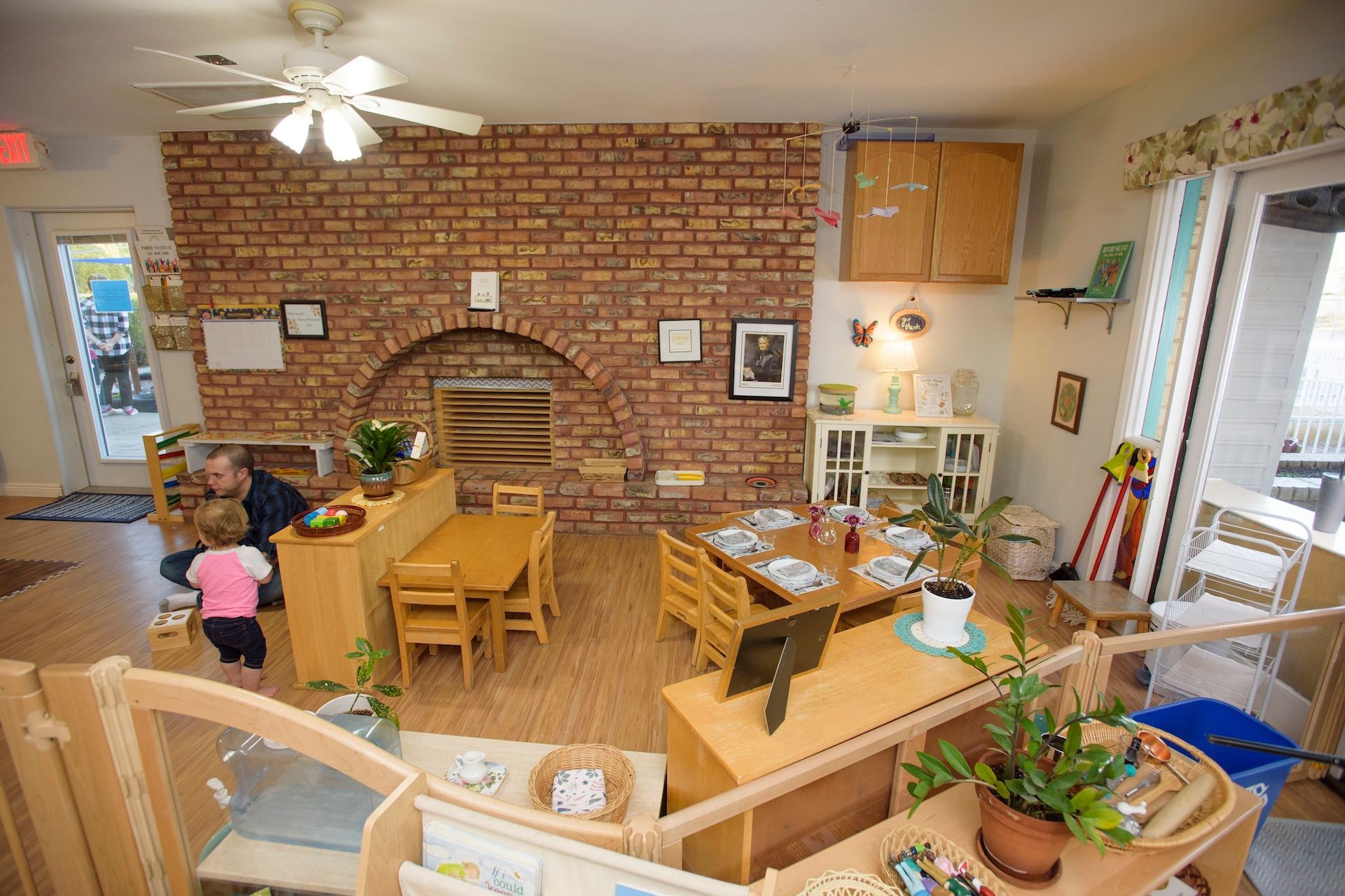 Childcare room with wooden tables, brick wall, and a child playing.