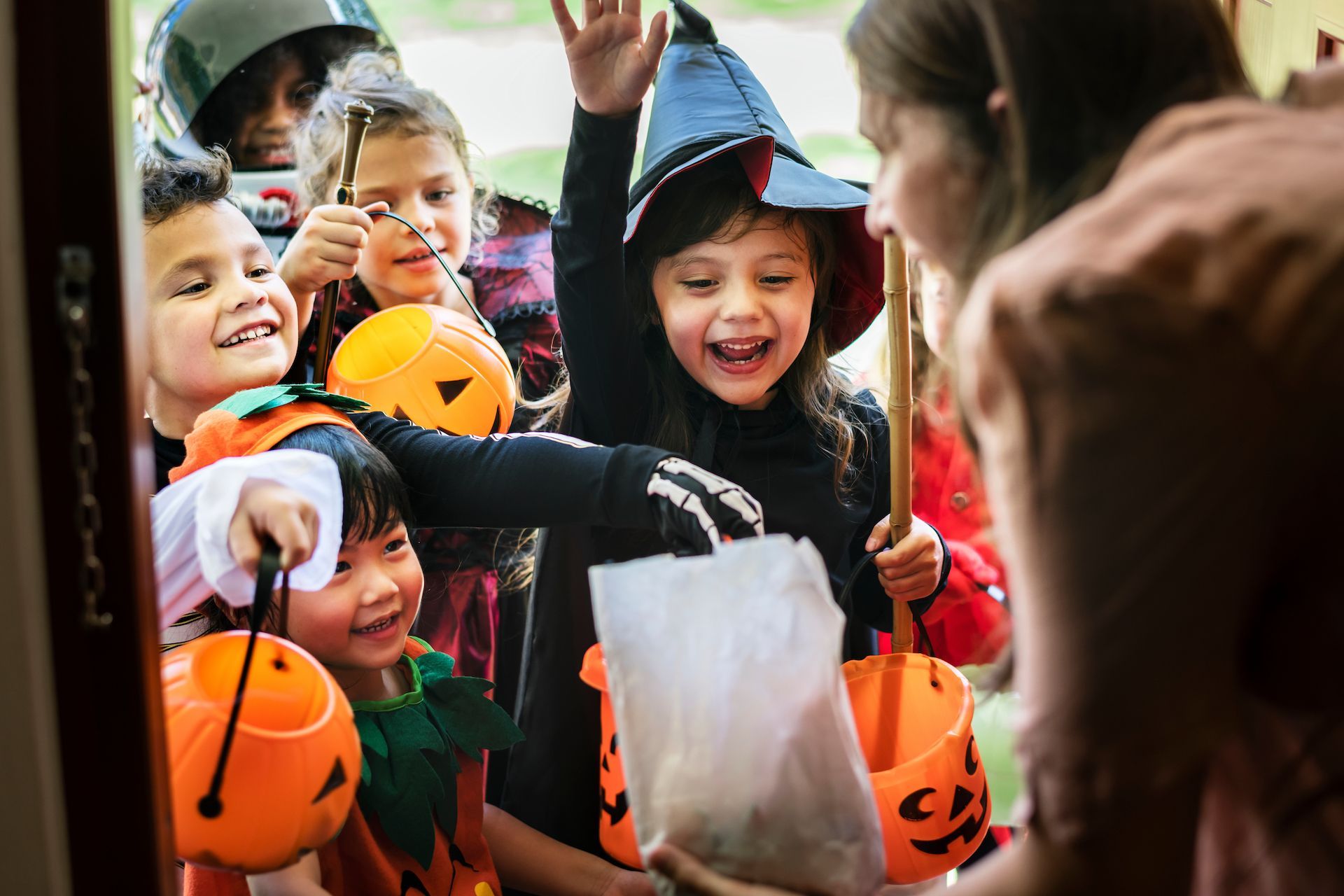 Children in Halloween costumes receive treats from a person at a doorway.