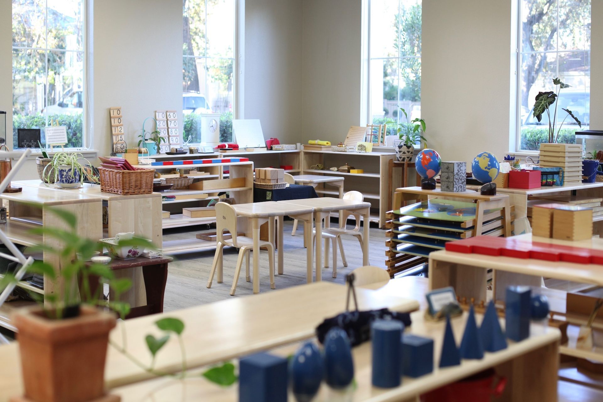 Montessori classroom with child-sized tables, shelves of educational materials, and large windows providing natural light.