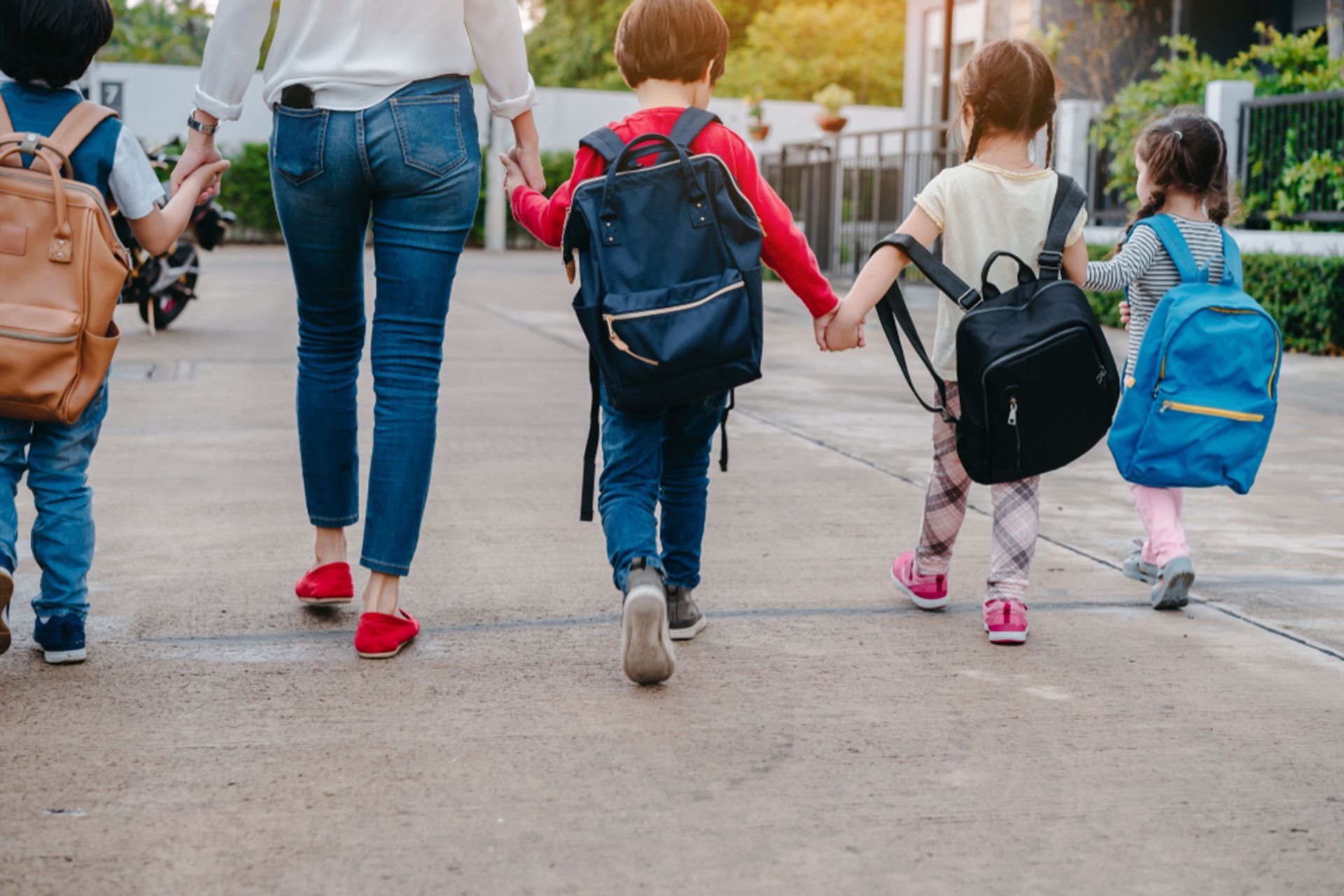 A woman and four children walk down a road, holding hands, wearing backpacks, on their way to school.