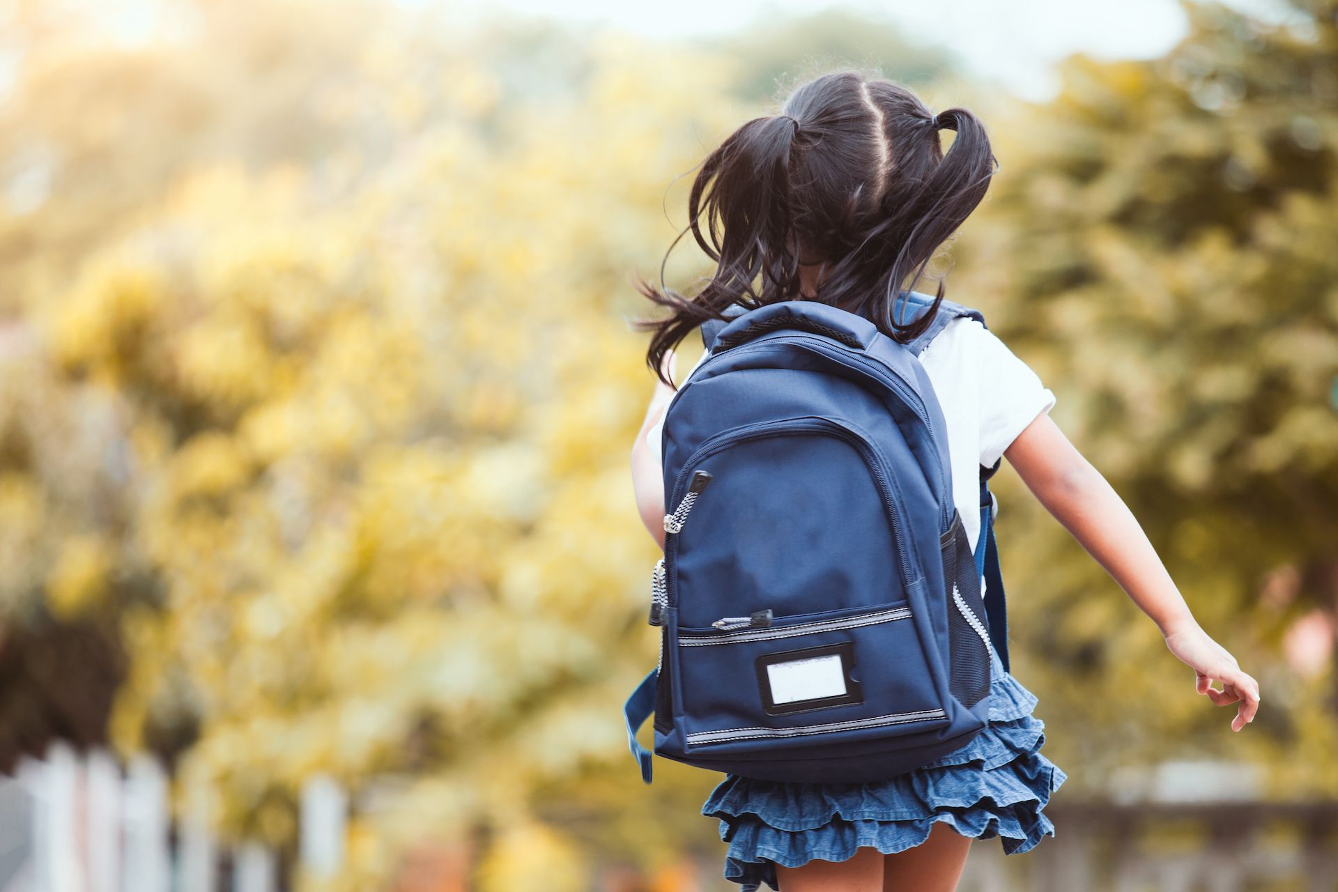 Girl with pigtails running away, wearing a blue backpack, outside near yellow and green trees.