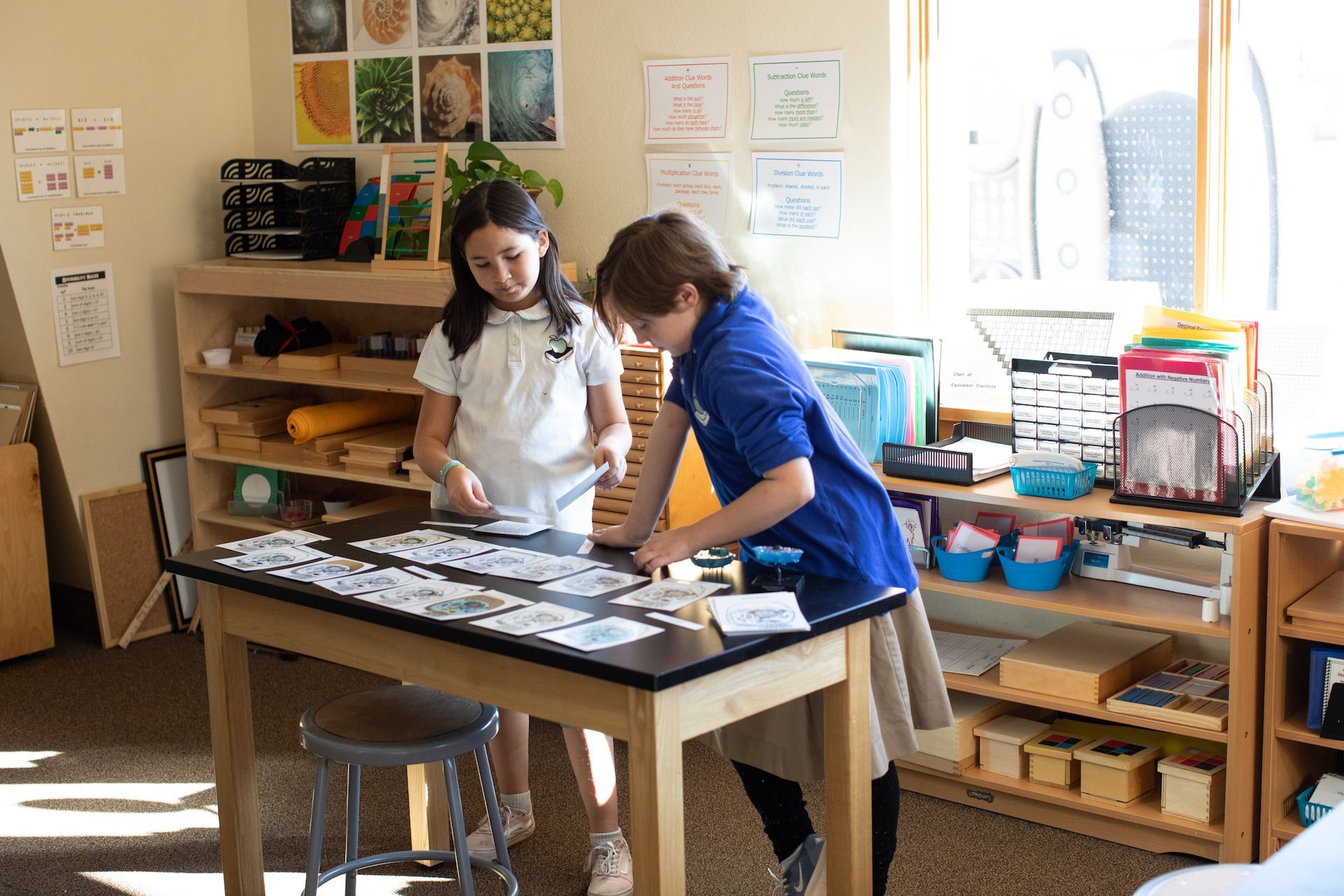 Two children at a table in a classroom sort cards. The room is bright with shelves and a window.