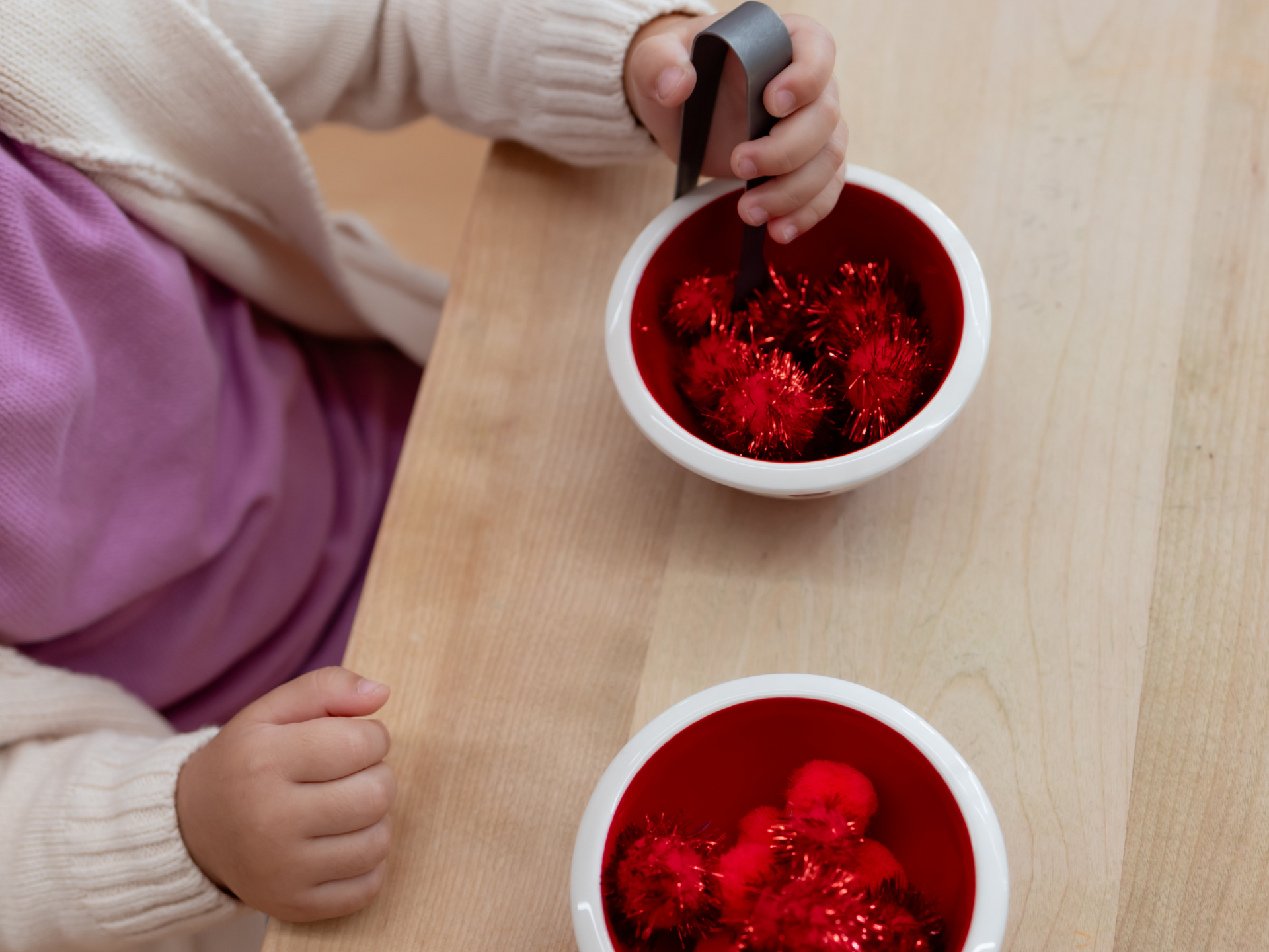 Child using tongs to transfer red objects from one bowl to another on a wooden table.