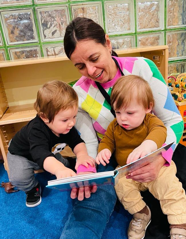 A Montessori guide is reading a book with two Montessori children. They are looking at the book, smiling.