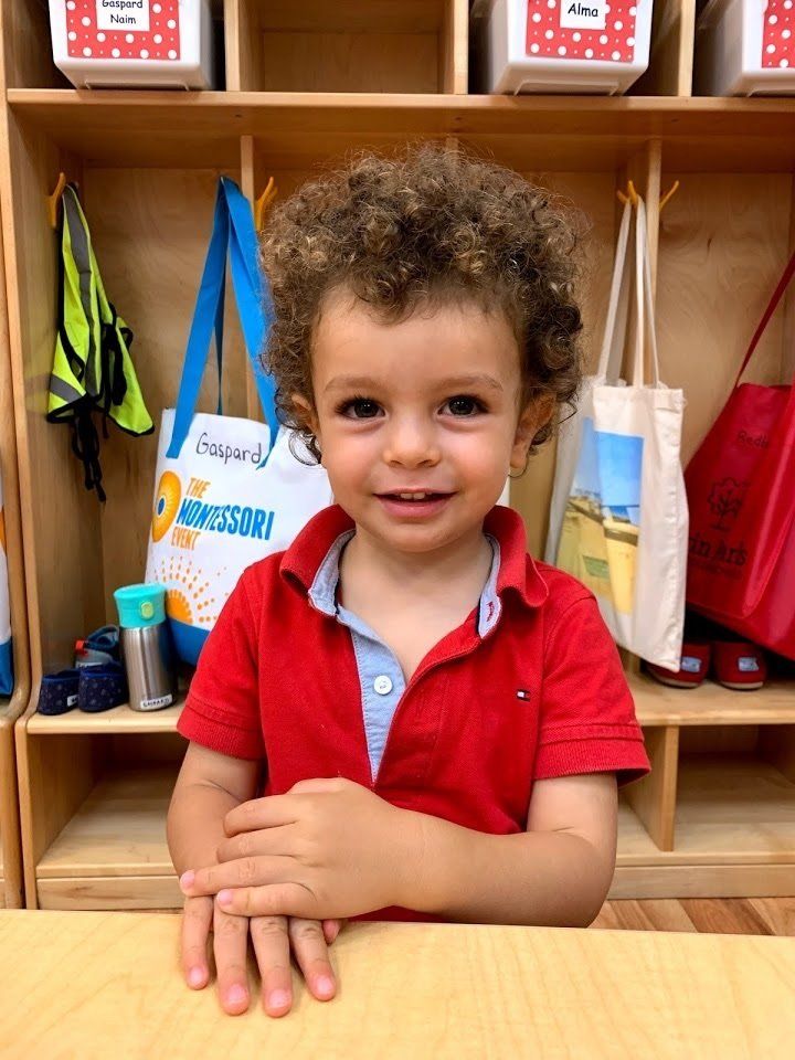 Young boy with curly hair smiles, wearing a red polo shirt, at a school cubby.
