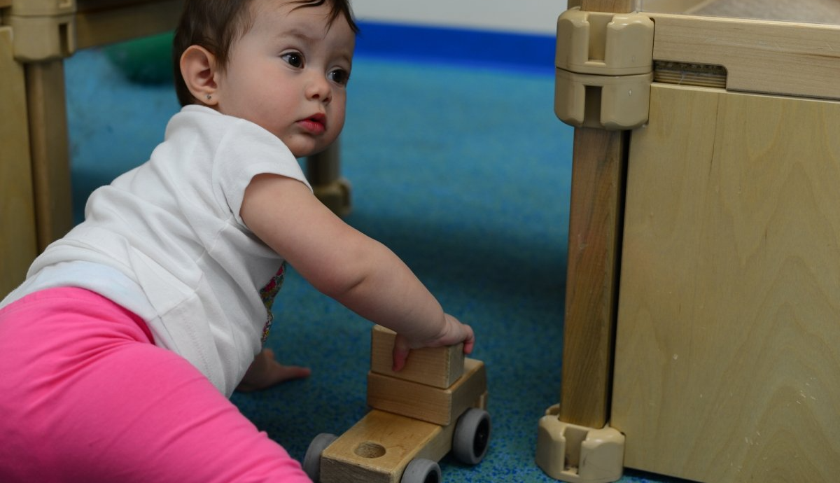 Baby reaching for wooden toy truck on blue carpet.