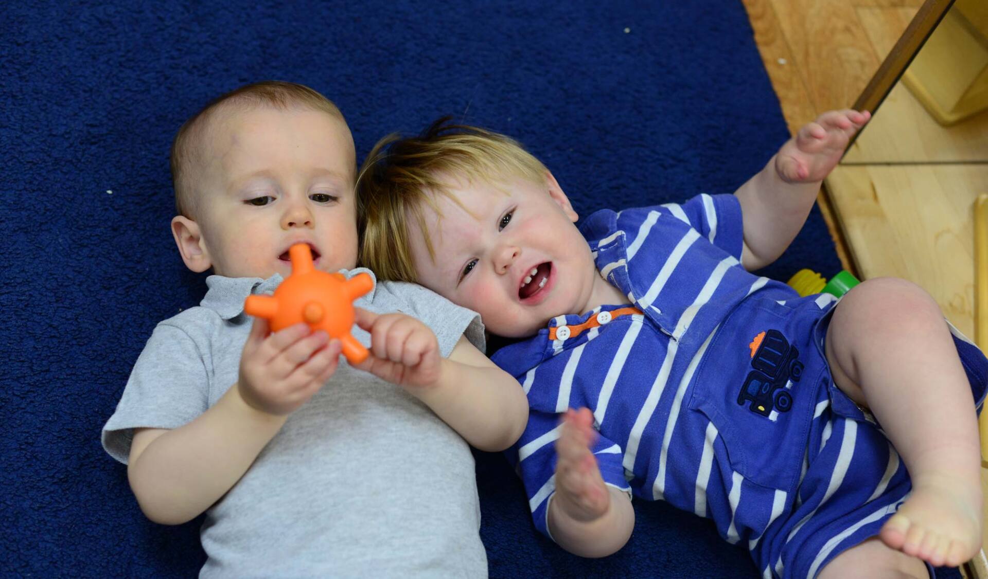 Two Montessori infants are on a blue carpet, one chewing a toy, the other laughing with arms up.