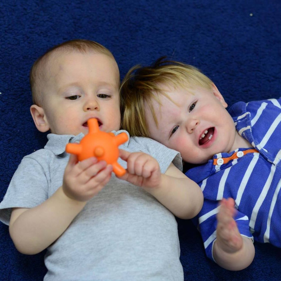 Two baby boys are laying on a blue carpet one is holding an orange toy in his mouth