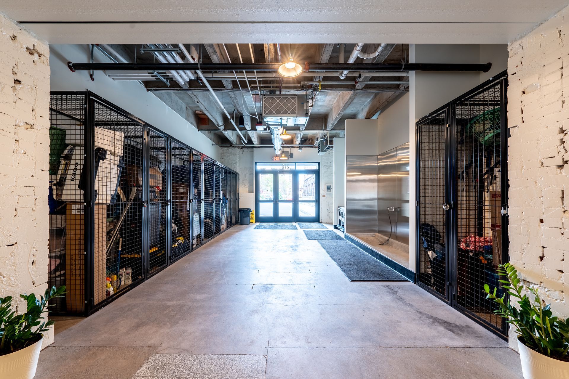 A hallway with storage lockers on both sides, leading to a doorway. Concrete floor, exposed ceiling with pipes and light fixtures at Zenith Apartments in Duluth, MN.