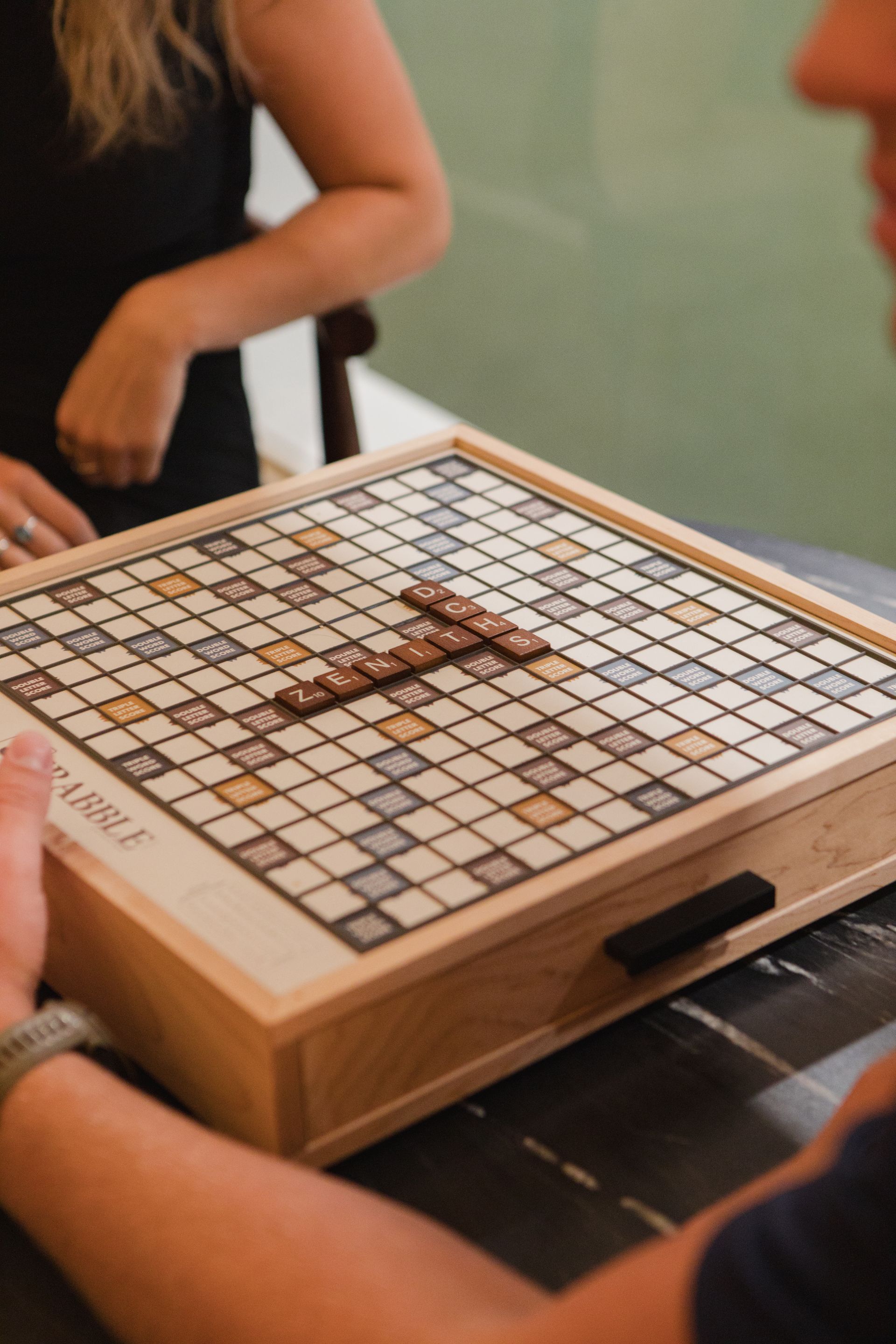 A person playing Scrabble on a wooden board. Another person is seated nearby at Zenith Apartments in Duluth, MN.