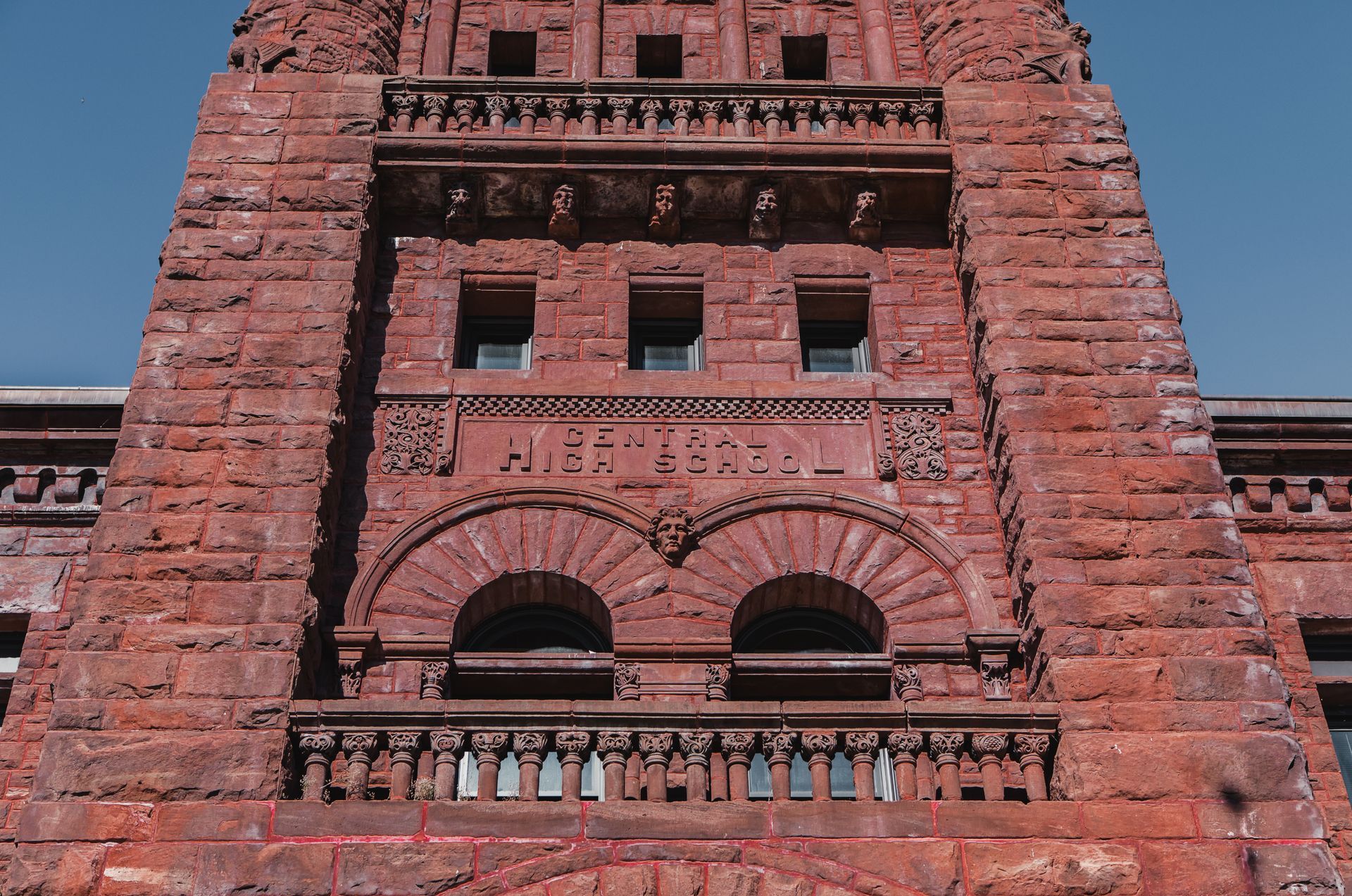 Red stone building facade with arched windows and a decorative balcony against blue sky at Zenith Apartments in Duluth, MN.