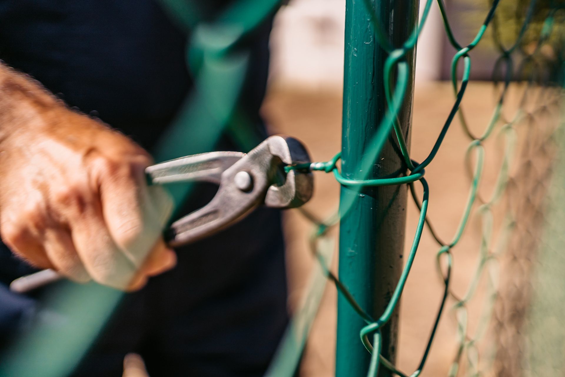 Close-up of a worker installing a green chain-link fence using pliers, demonstrating professional fence installation and repair services by Mobile Fence Co. in Daphne, AL. Close-up of a worker installing a green chain-link fence using pliers, demonstrating professional fence installation and repair services by Mobile Fence Co. in Daphne, AL.