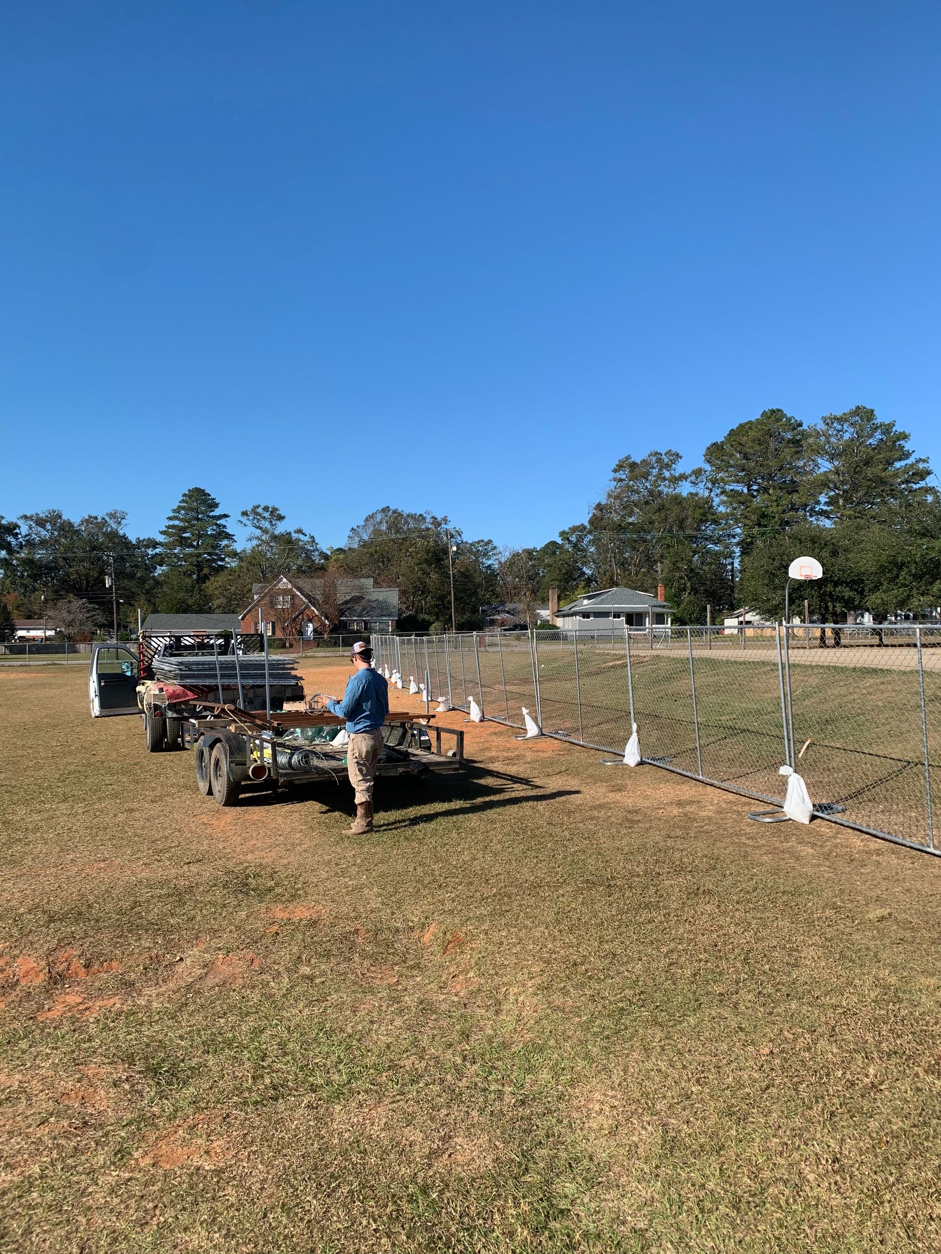 Person standing near a tractor in a grassy field. Clear blue sky, trees, and fence visible.