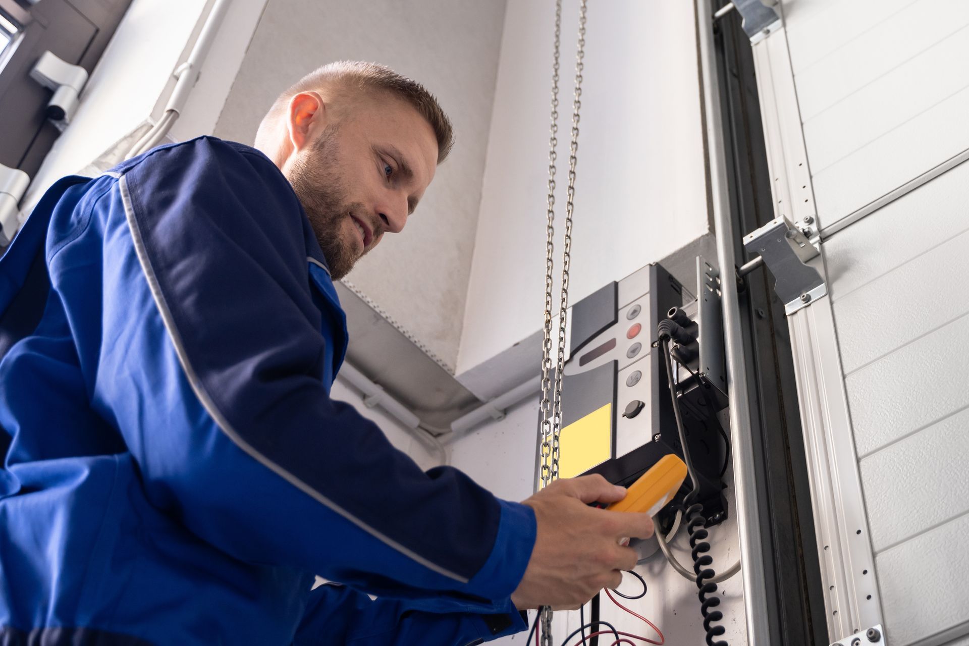 Man in blue coveralls checks control panel with a yellow multimeter