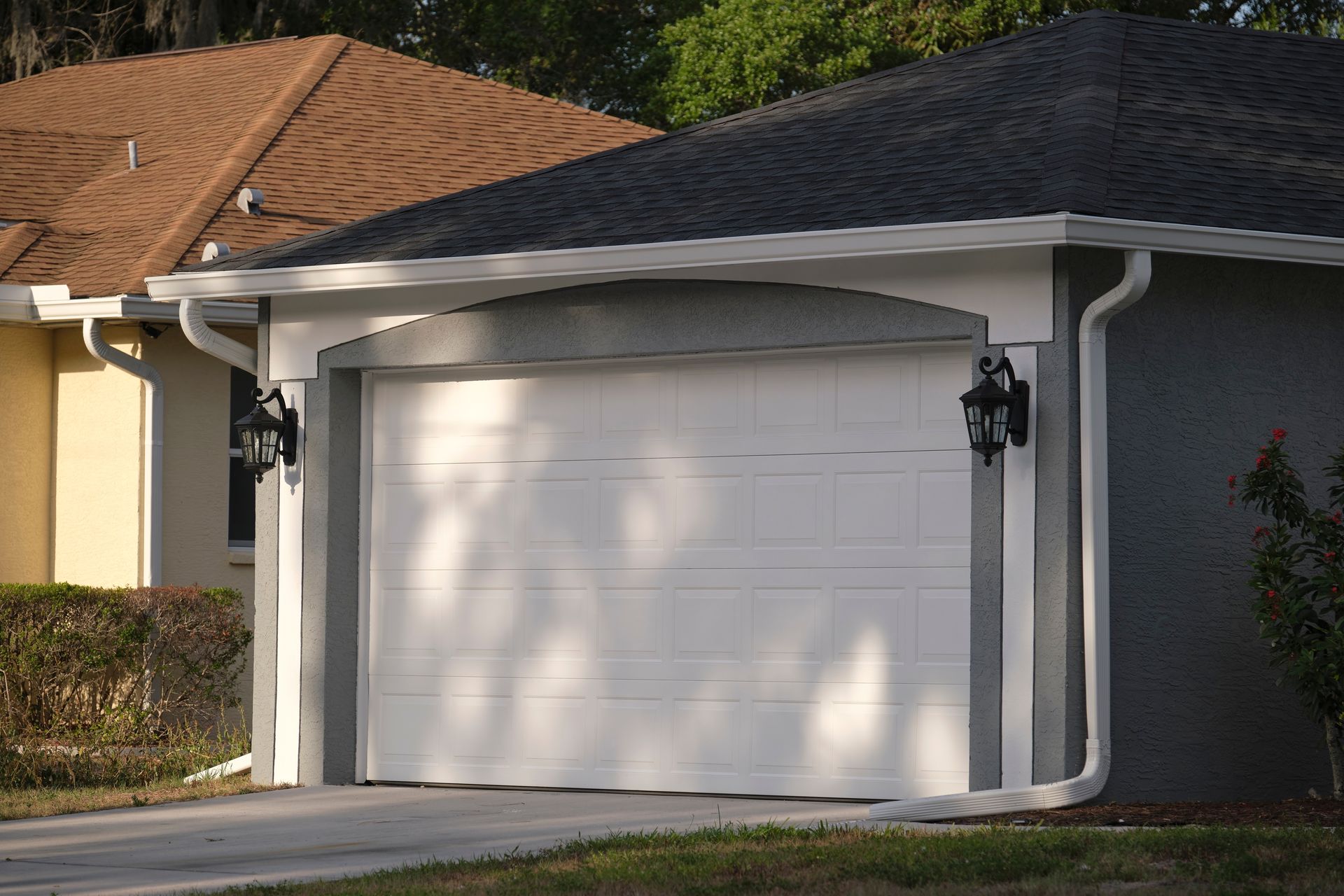 White garage door on a house with gray siding and black wall lanterns in a suburban driveway.