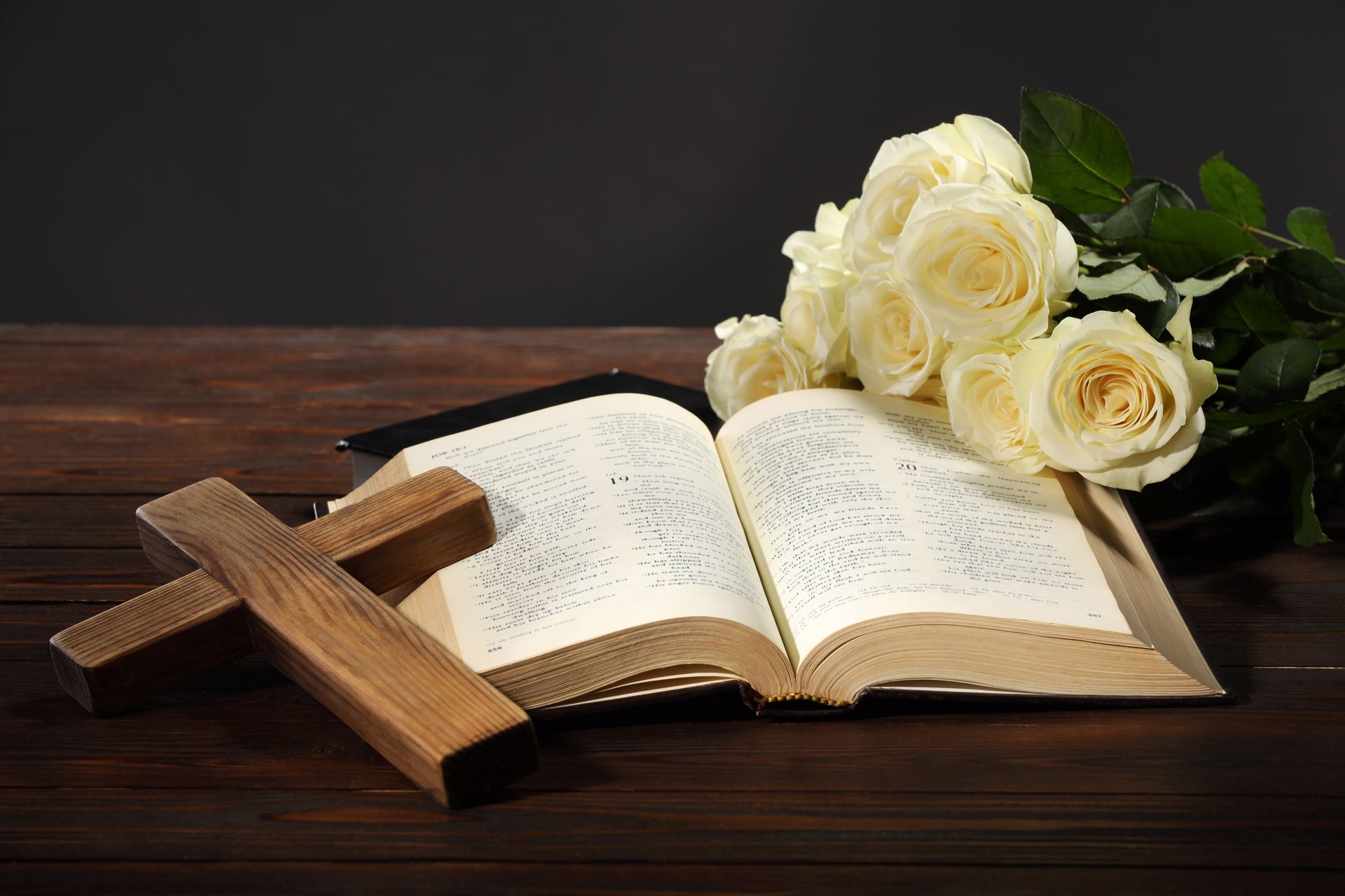 Open Bible with wooden cross and white roses on a dark wooden table