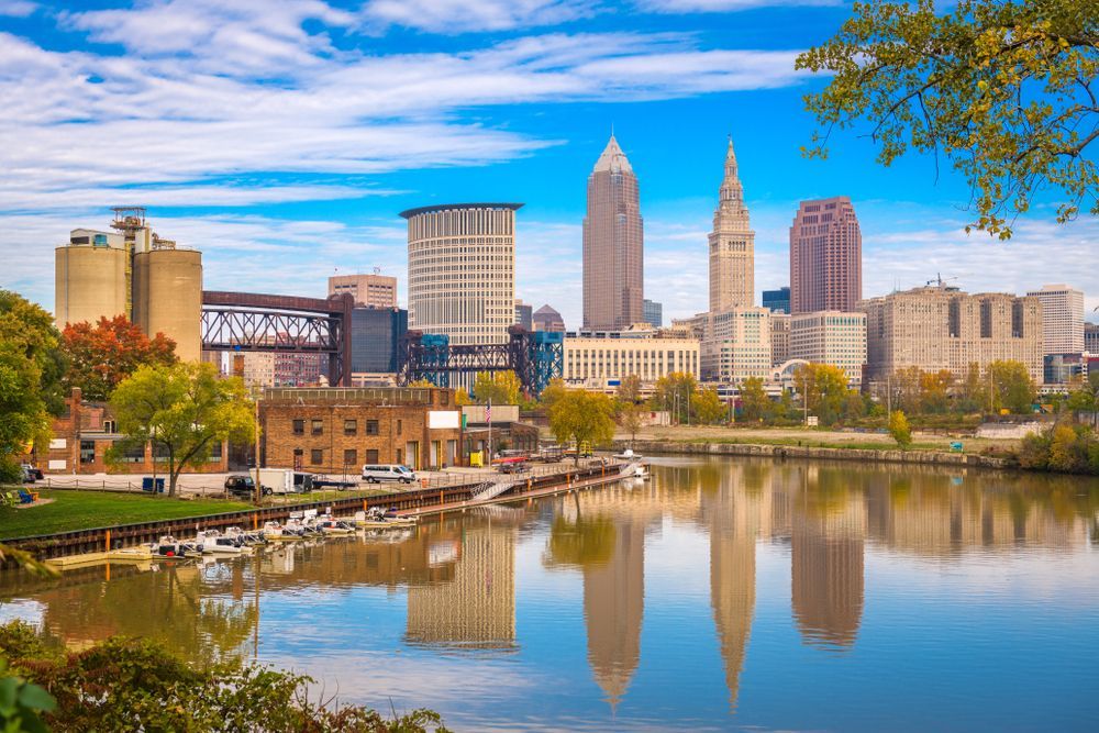 Cleveland skyline reflected in a calm river under a blue sky with autumn trees in the foreground
