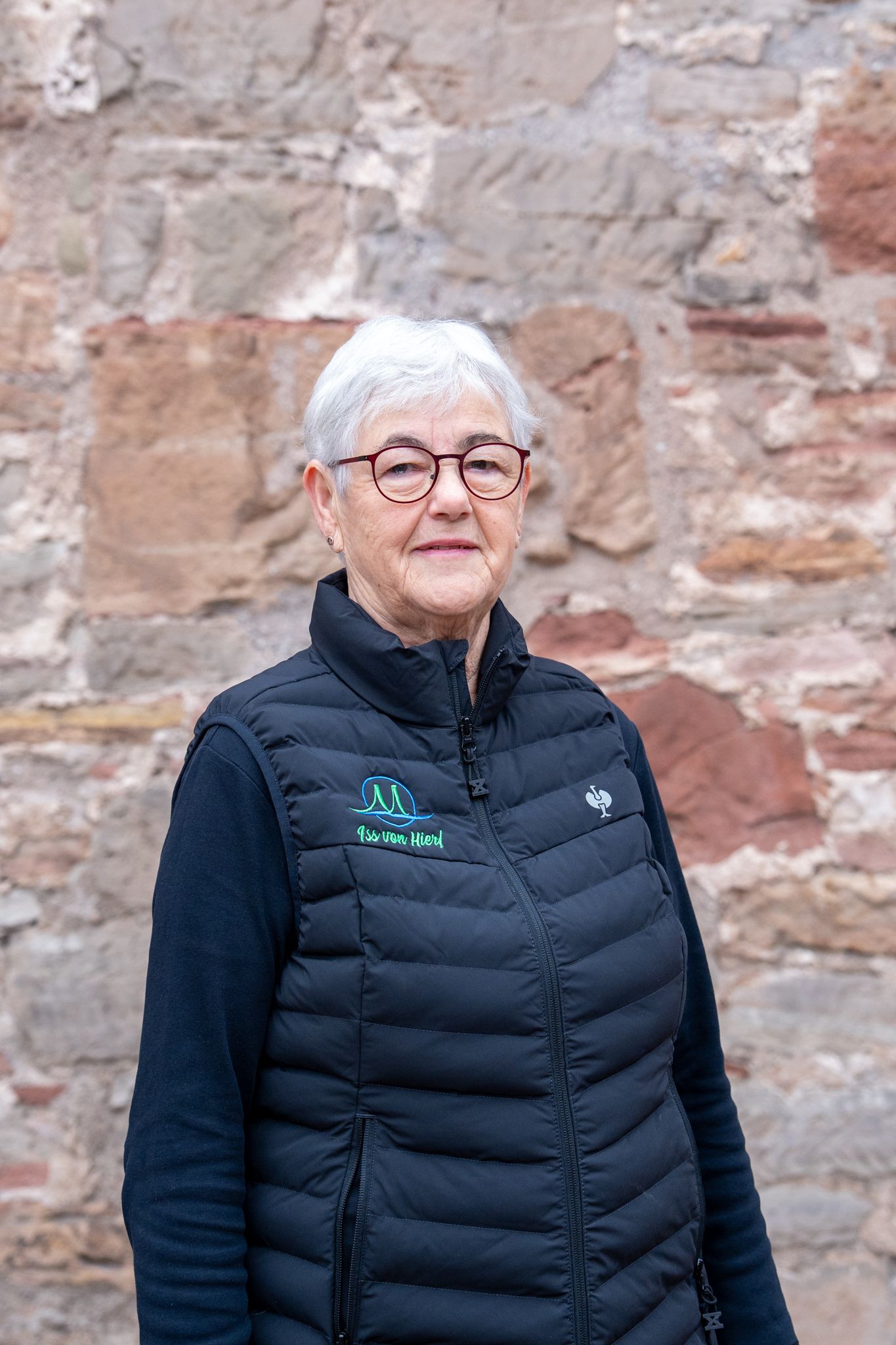Eine Frau mit Brille und Weste lächelt vor einer Steinmauer.