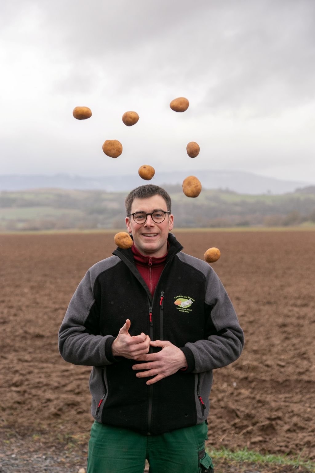Ein Mann jongliert auf einem Feld mit Kartoffeln. Er trägt eine Jacke, eine Brille und lächelt; im Hintergrund sind bewölkte Hügel zu sehen.
