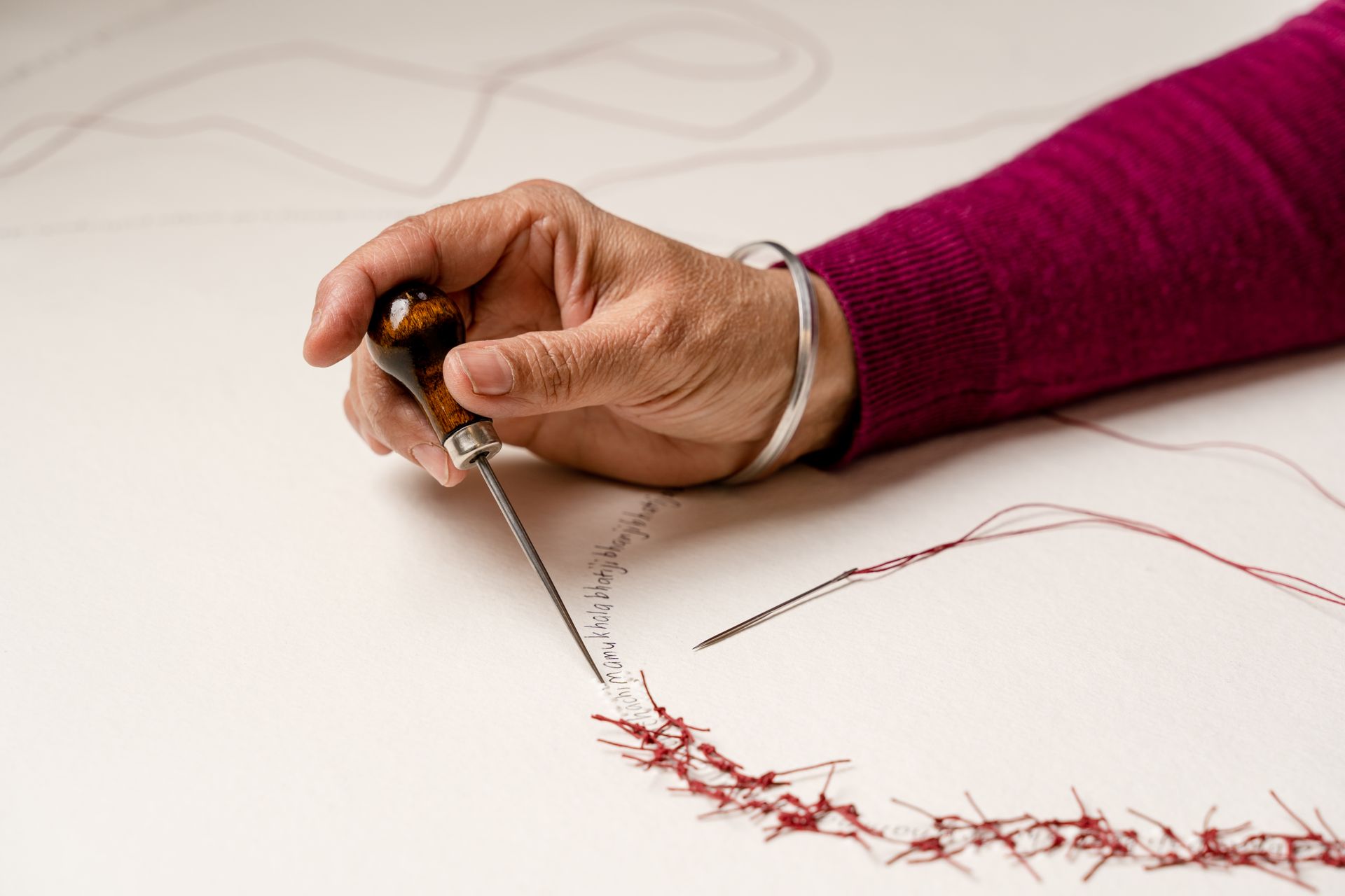 Close-up of hand holding a tool, stitching red thread onto white fabric. A sleeve in magenta is visible.