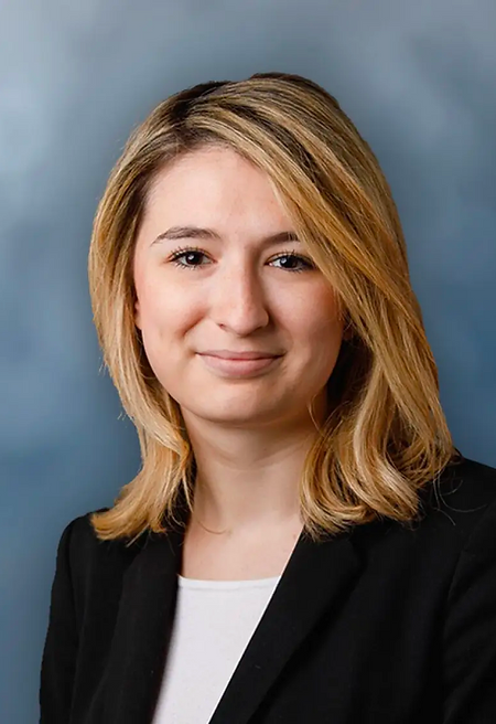 Headshot of a person in a black blazer and white top against a blue-gray background