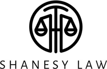 Black circular law firm logo with scales of justice and the text “SHANESY LAW” below.