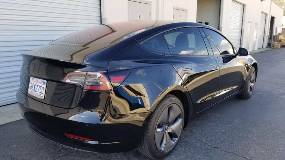 Black Tesla sedan parked beside a white industrial building in a lot.