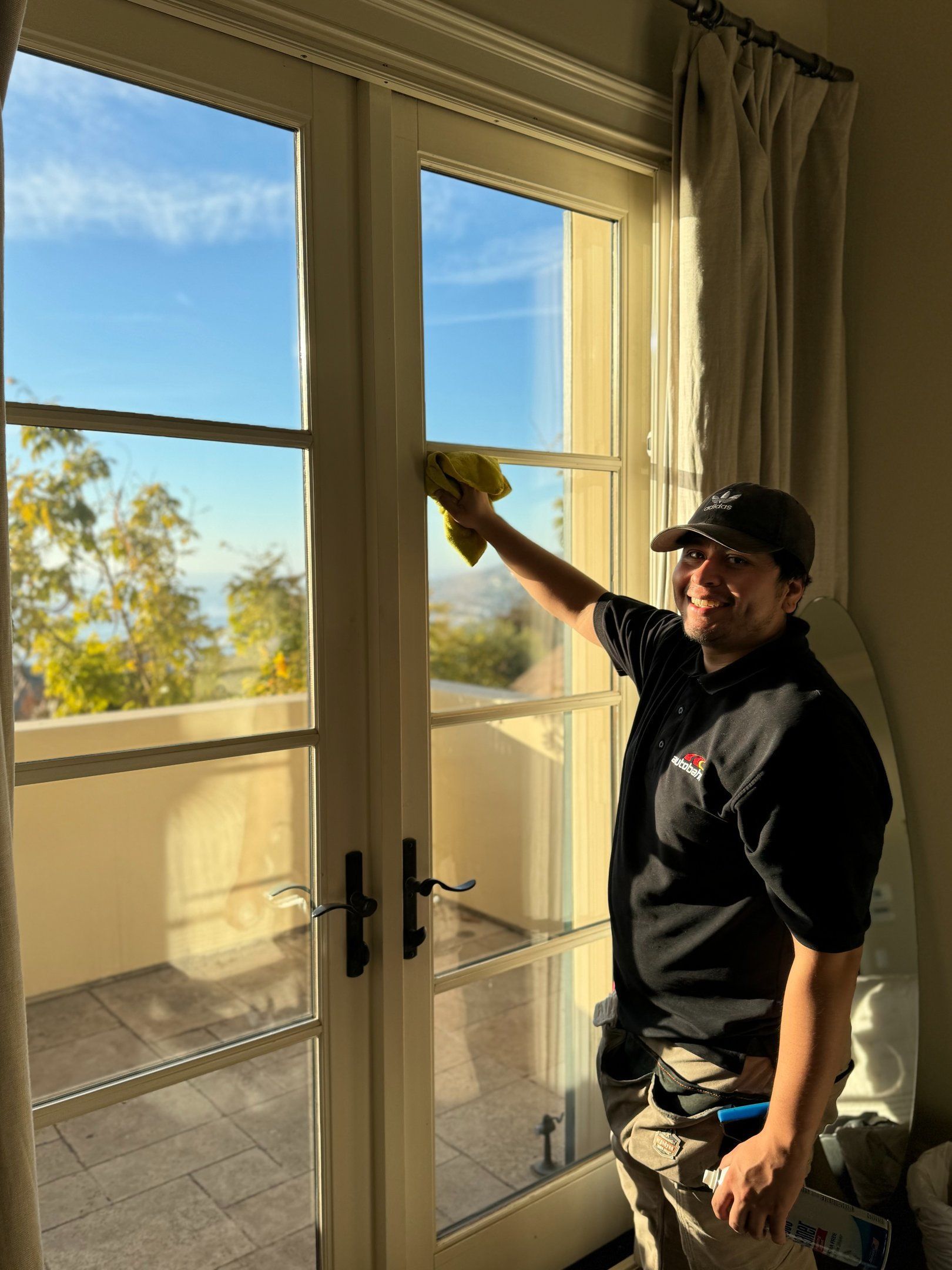Person wiping a tall glass door indoors, sunlight streaming in from outside.