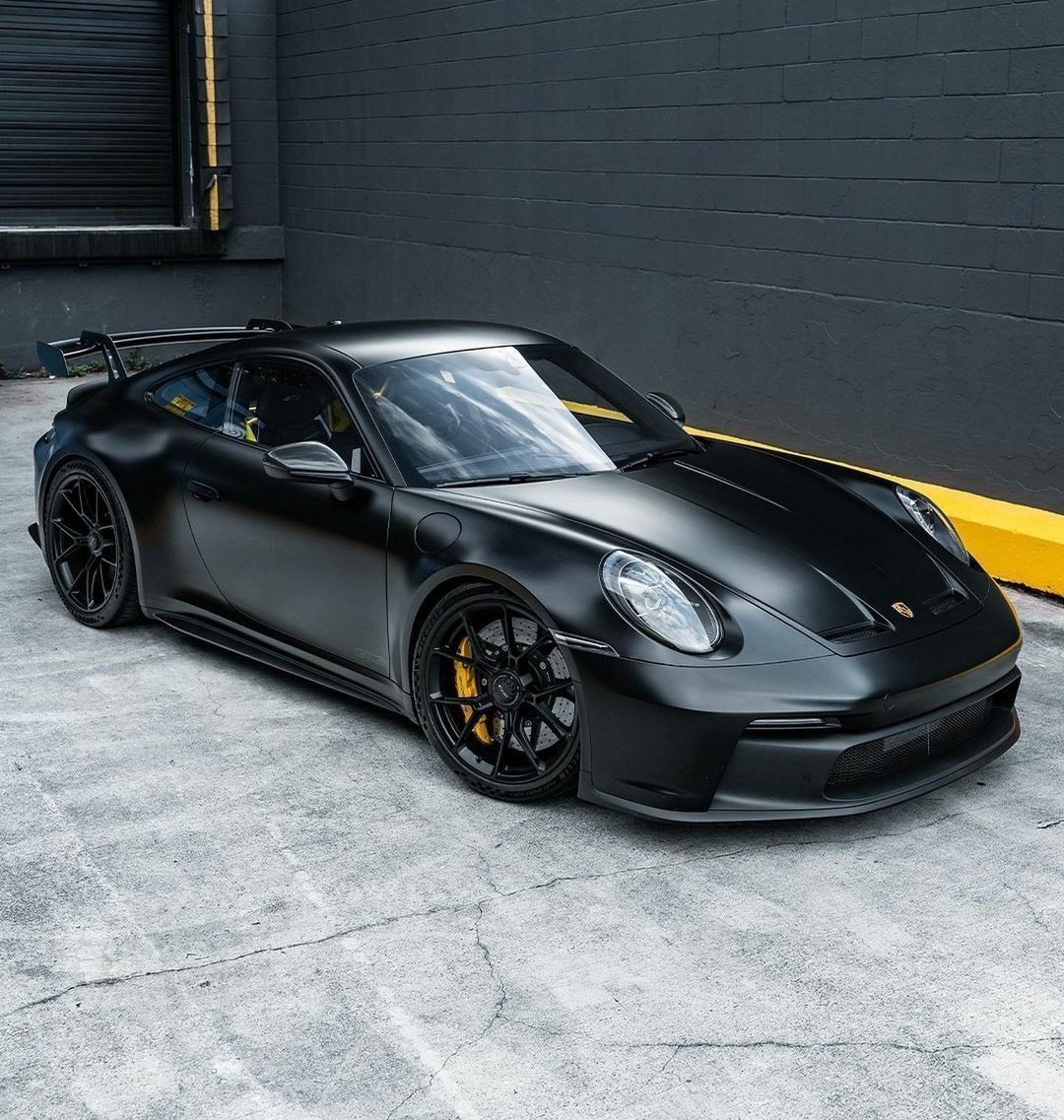 Matte black sports car with yellow accents parked beside a dark brick wall.