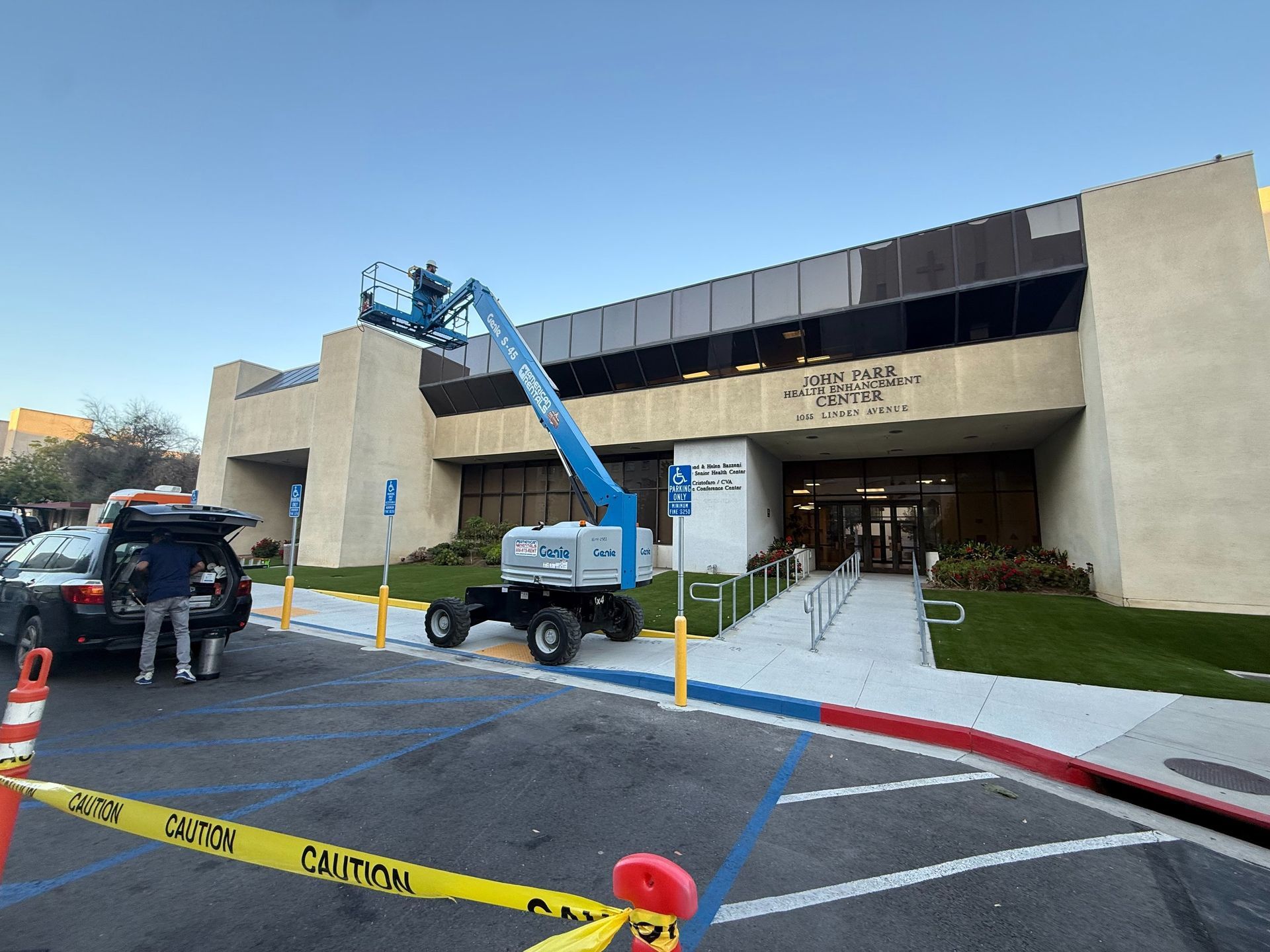 Construction site with a blue lift in front of a modern building entrance, bordered by caution tape.